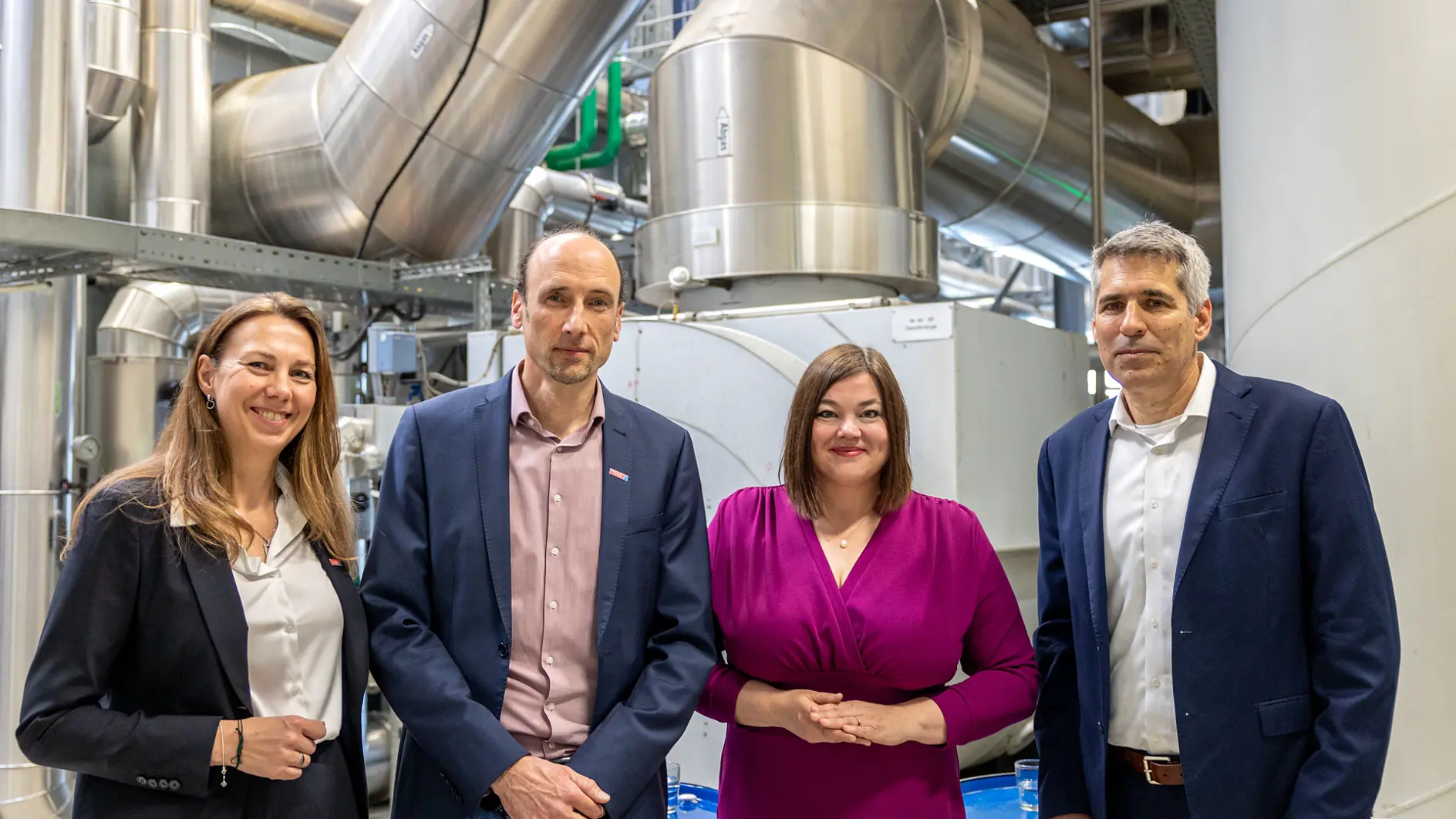 Four people in business attire stand before large machinery and pipes, posing for a group photo at a tesa tape industrial facility.