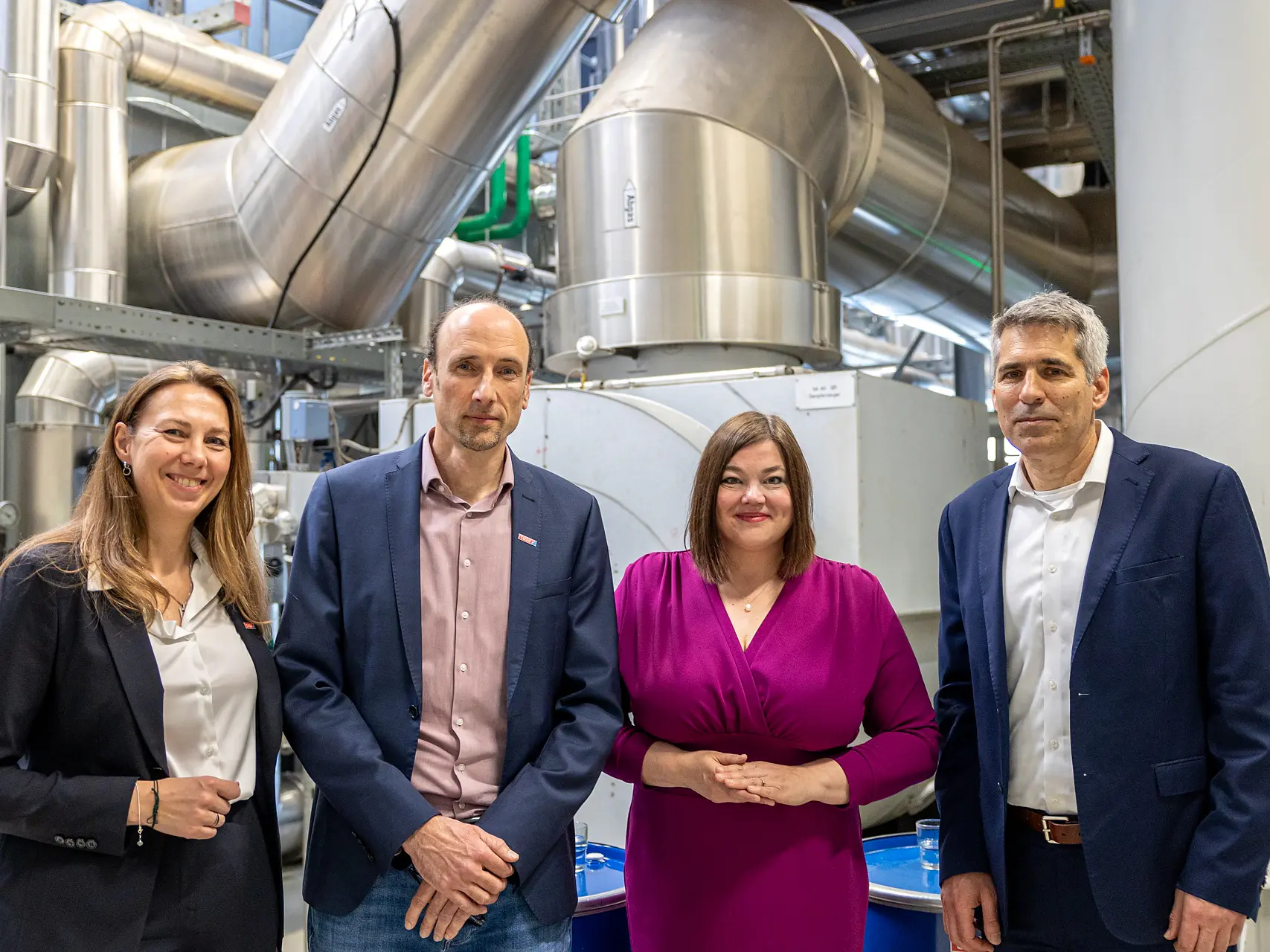 Four people in business attire stand before large machinery and pipes, posing for a group photo at a tesa tape industrial facility. (This text has been generated by AI)