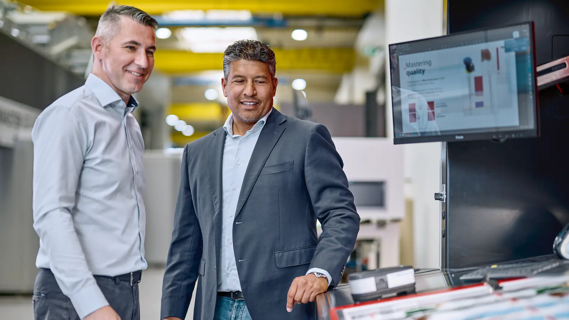 Two men stand at a workstation with a monitor and printed materials, smiling as they discuss something on the screen, featuring tesa tape. (This text has been generated by AI)