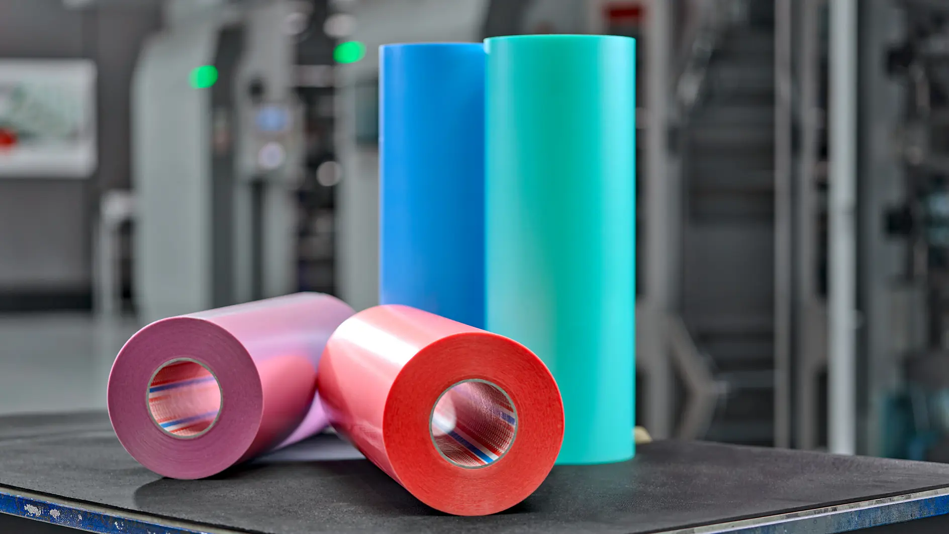 Five rolls of colored adhesive tape in blue, teal, pink, red, and gray on a metal table in an industrial setting. (This text has been generated by AI)