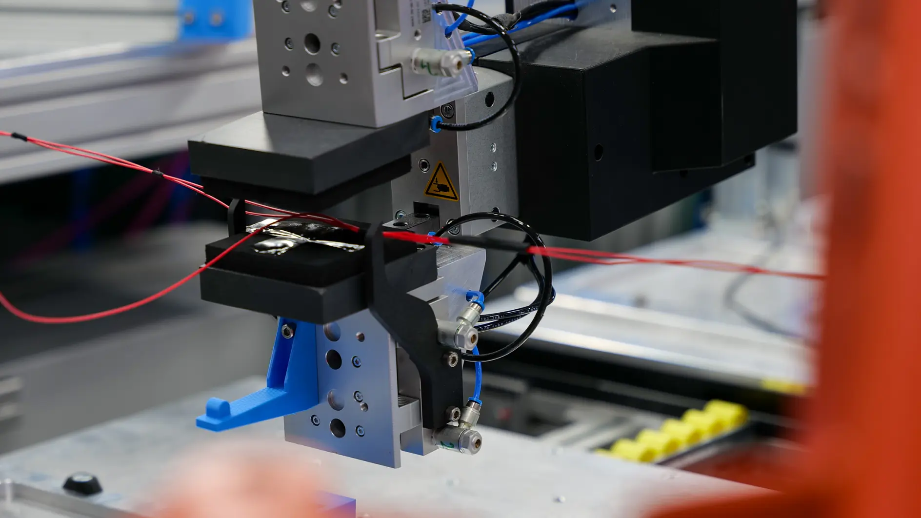 A close-up of an industrial robotic arm with clamps handling red wires secured by tesa tape, set in a factory with machinery.