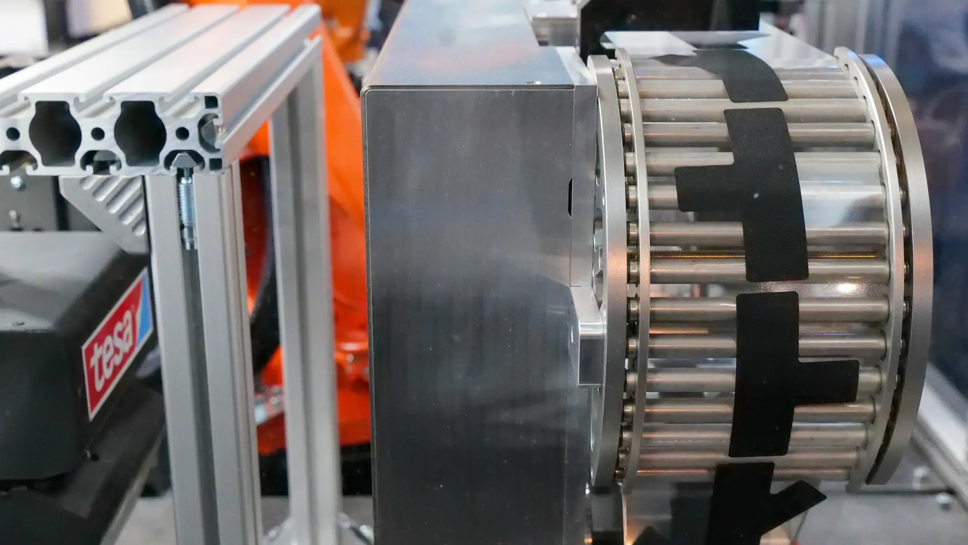 A close-up of industrial machinery featuring a metal cylinder with tesa tape, an orange robotic arm behind, and a frame labeled tese.