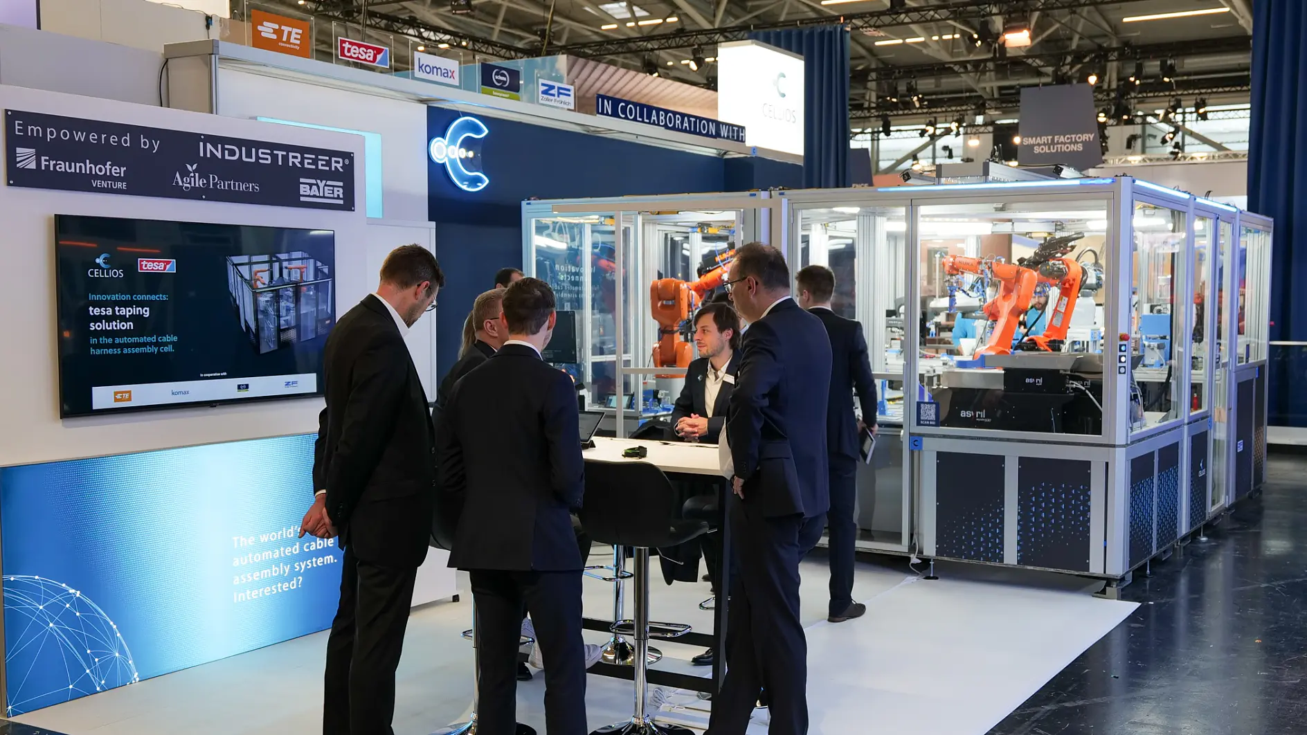 Five people in business attire meet at a trade show booth featuring tesa tape, with machinery, robots, and tech partner info screens.