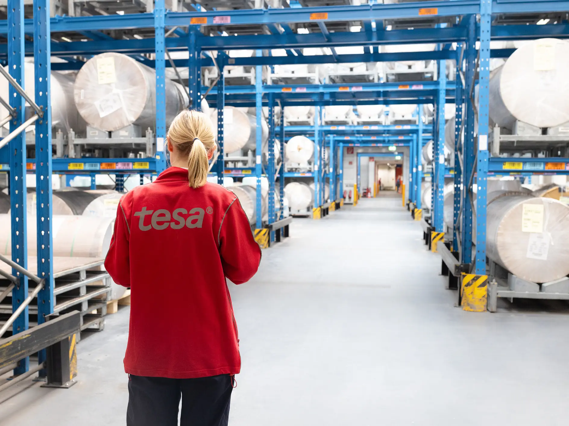 A person in a red jacket with tesa on the back stands in a warehouse aisle, surrounded by large rolls of tesa tape on blue shelves. (This text has been generated by AI)