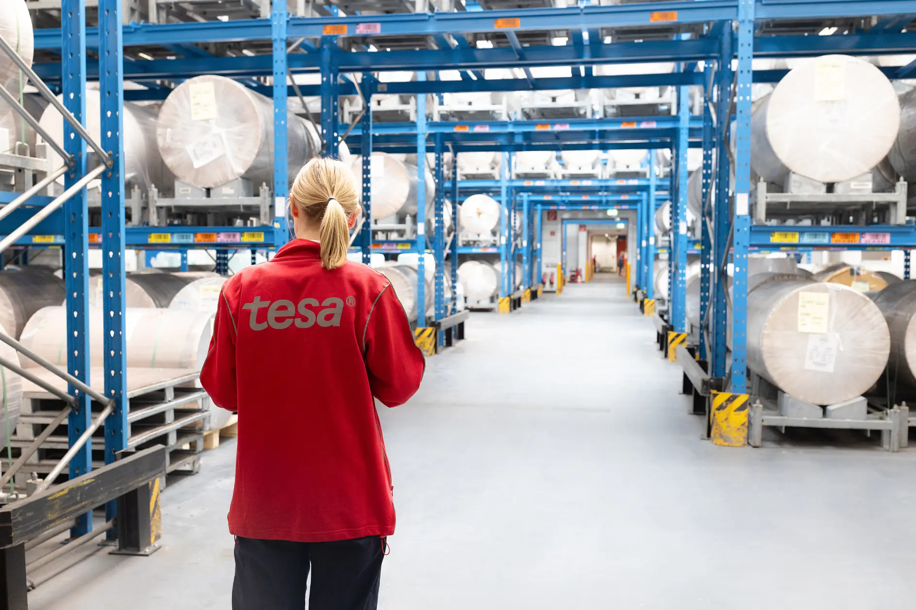 A person in a red jacket with tesa on the back stands in a warehouse aisle, surrounded by large rolls of tesa tape on blue shelves. (This text has been generated by AI)