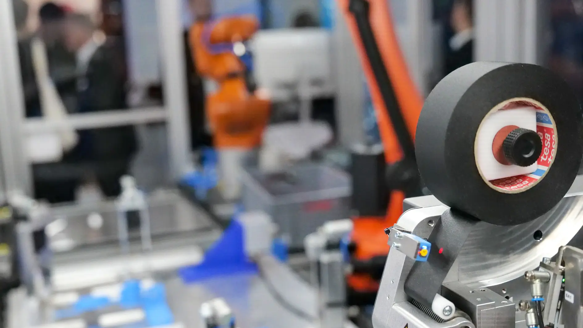 Close-up of industrial robotic machinery applying tesa tape, with an orange robotic arm and equipment in a manufacturing or lab setting.