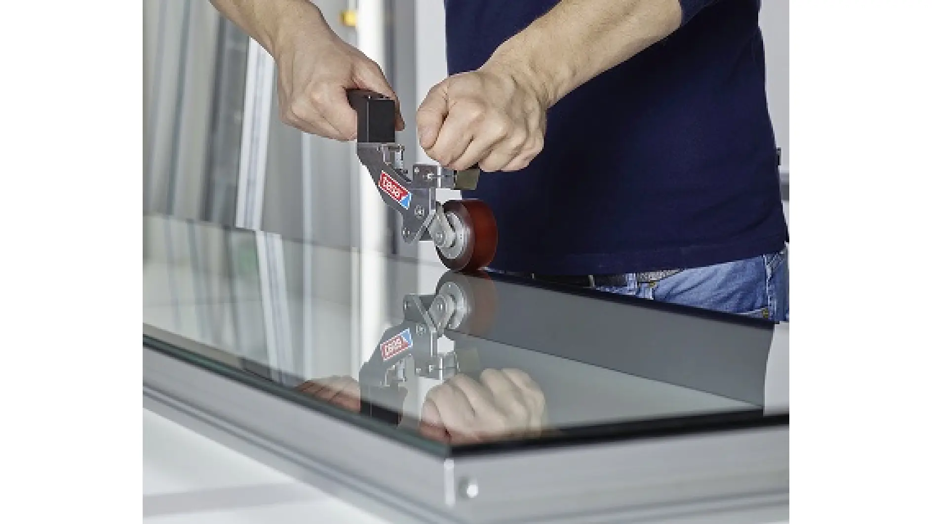 A person uses a handheld tool with a red wheel to work on the edge of a glass panel mounted in a metal frame with tesa tape. (This text has been generated by AI)