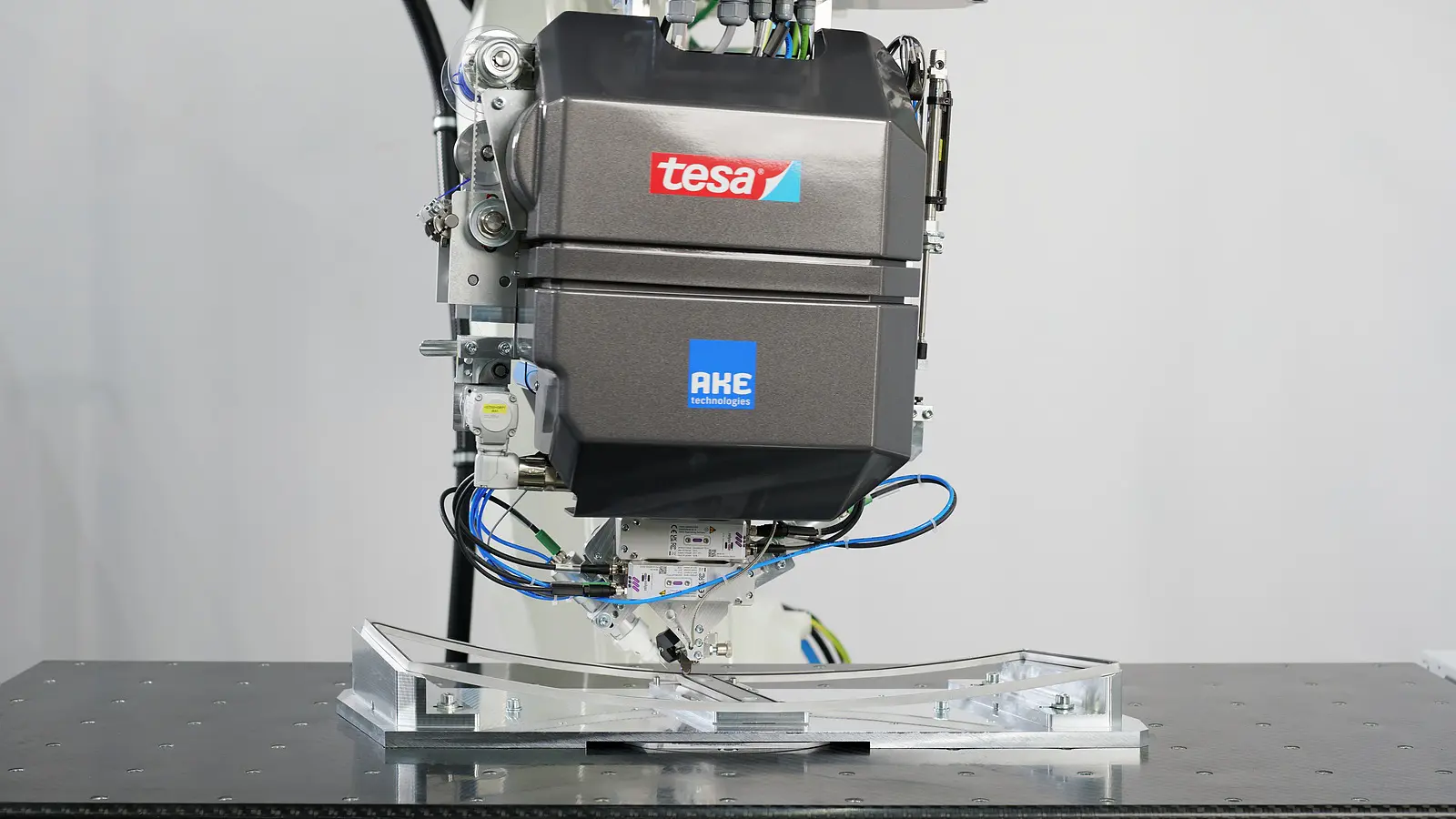 A close-up of a robotic machine with tesa tape and AKE logos, wires, and mechanical parts above a metallic fixture on a black table.