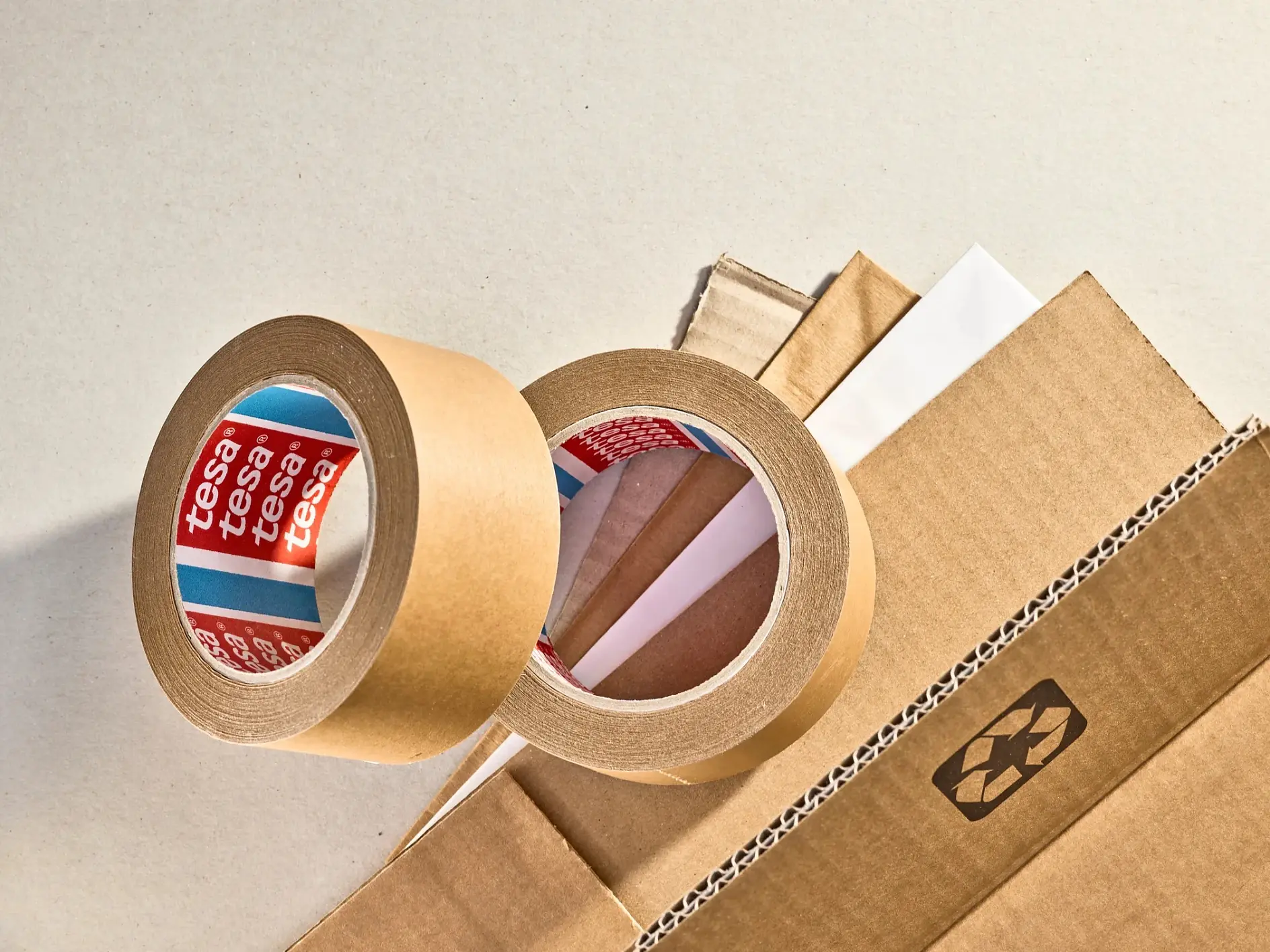 Two rolls of brown tesa tape sit beside corrugated cardboard pieces with some white paper layers, labels in red and blue. Well-lit scene. (This text has been generated by AI)