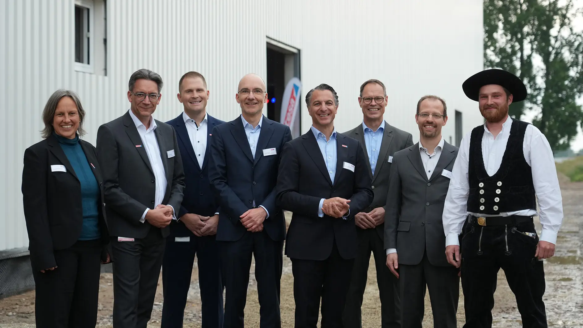 Eight people in business attire and one in workwear stand outside a white industrial building, smiling; gravel ground, trees behind. (This text has been generated by AI)