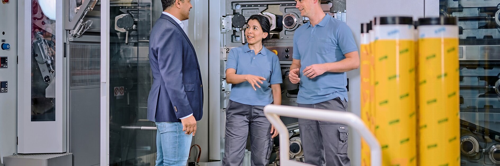 Three people in a factory setting examine large rolls of yellow tesa adhesive tape, surrounded by industrial machinery and equipment.