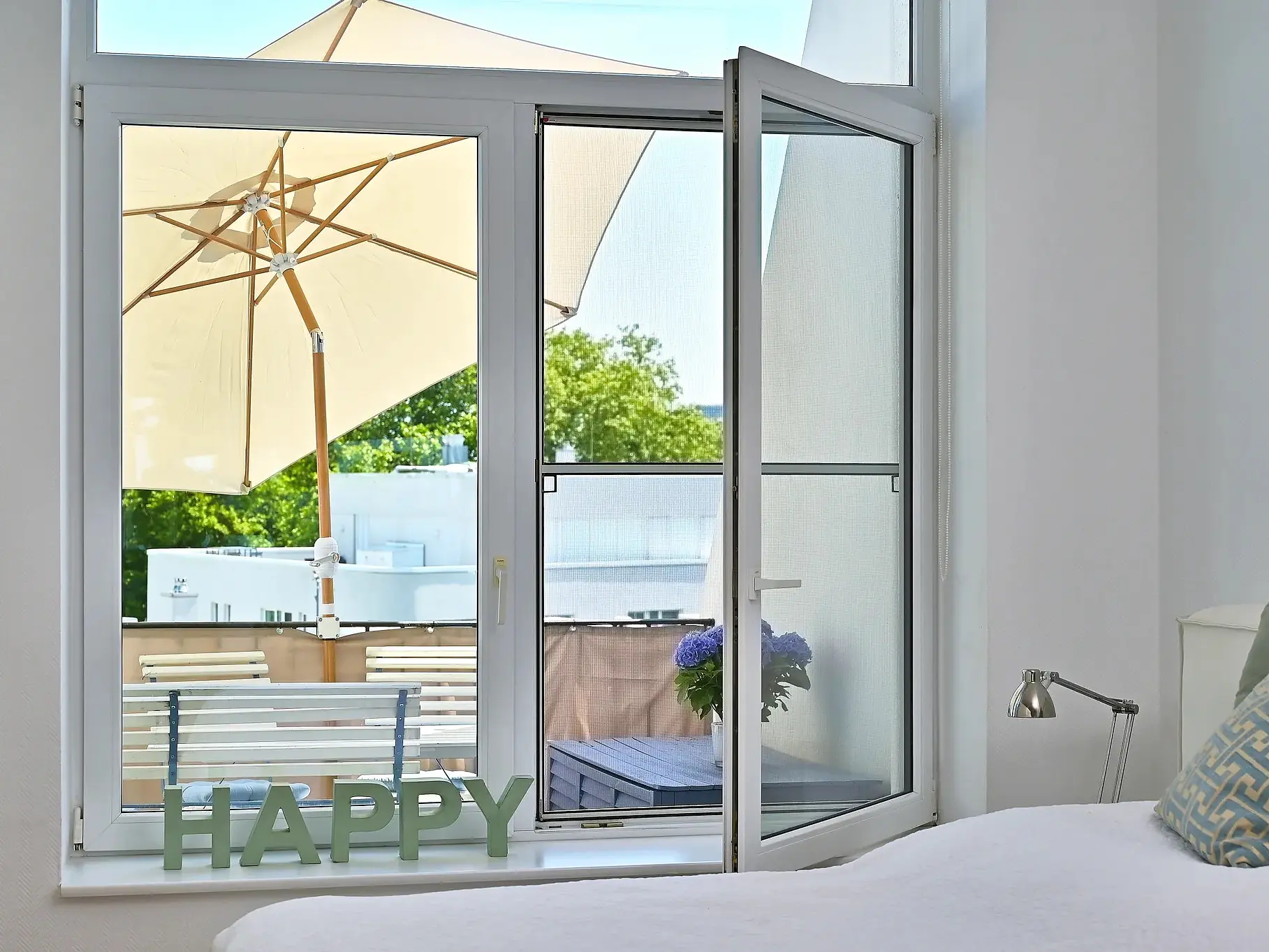 A bedroom with a bed, silver reading lamp, and decorative letters spelling HAPPY. An open window shows a patio with a large umbrella, outdoor furniture, and greenery outside. (This text has been generated by AI)