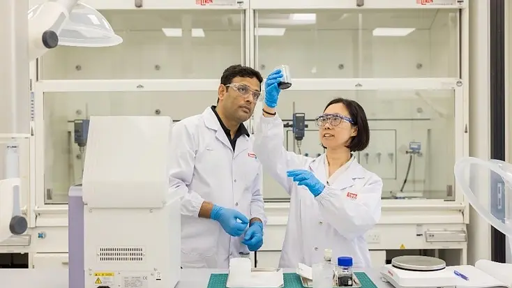 Two scientists wearing lab coats, gloves, and safety glasses examine a sample in a laboratory. One holds the sample up with tesa tape applied, while the other observes. Laboratory equipment is visible on the table and in the background.