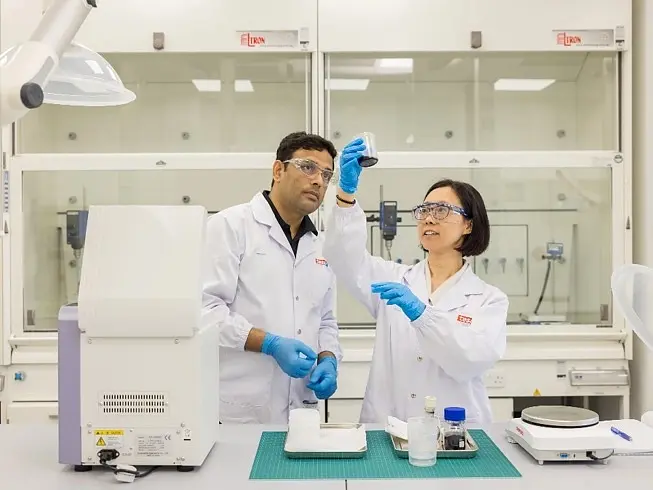 Two scientists wearing lab coats, gloves, and safety glasses examine a sample in a laboratory. One holds the sample up with tesa tape applied, while the other observes. Laboratory equipment is visible on the table and in the background. (This text has been generated by AI)