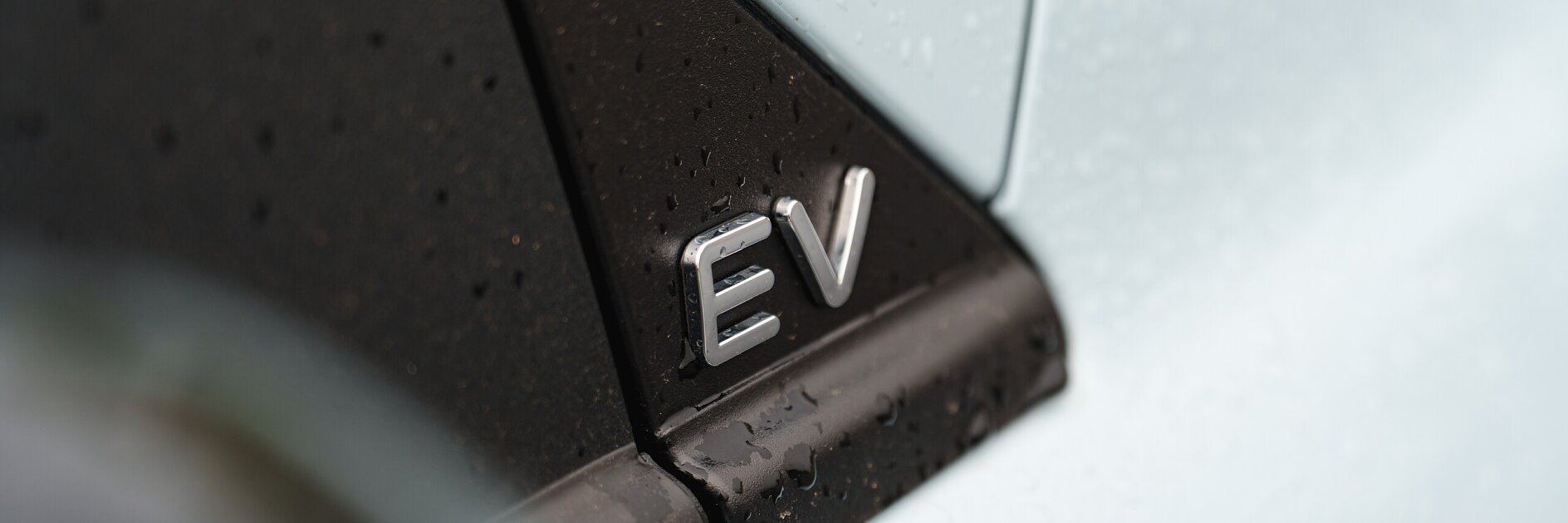 Close-up of a car with water droplets on its surface, showing a silver EV badge on a black panel, indicating the vehicle is electric. A strip of tesa tape is visible adjacent to the badge. The paint appears light blue or white.