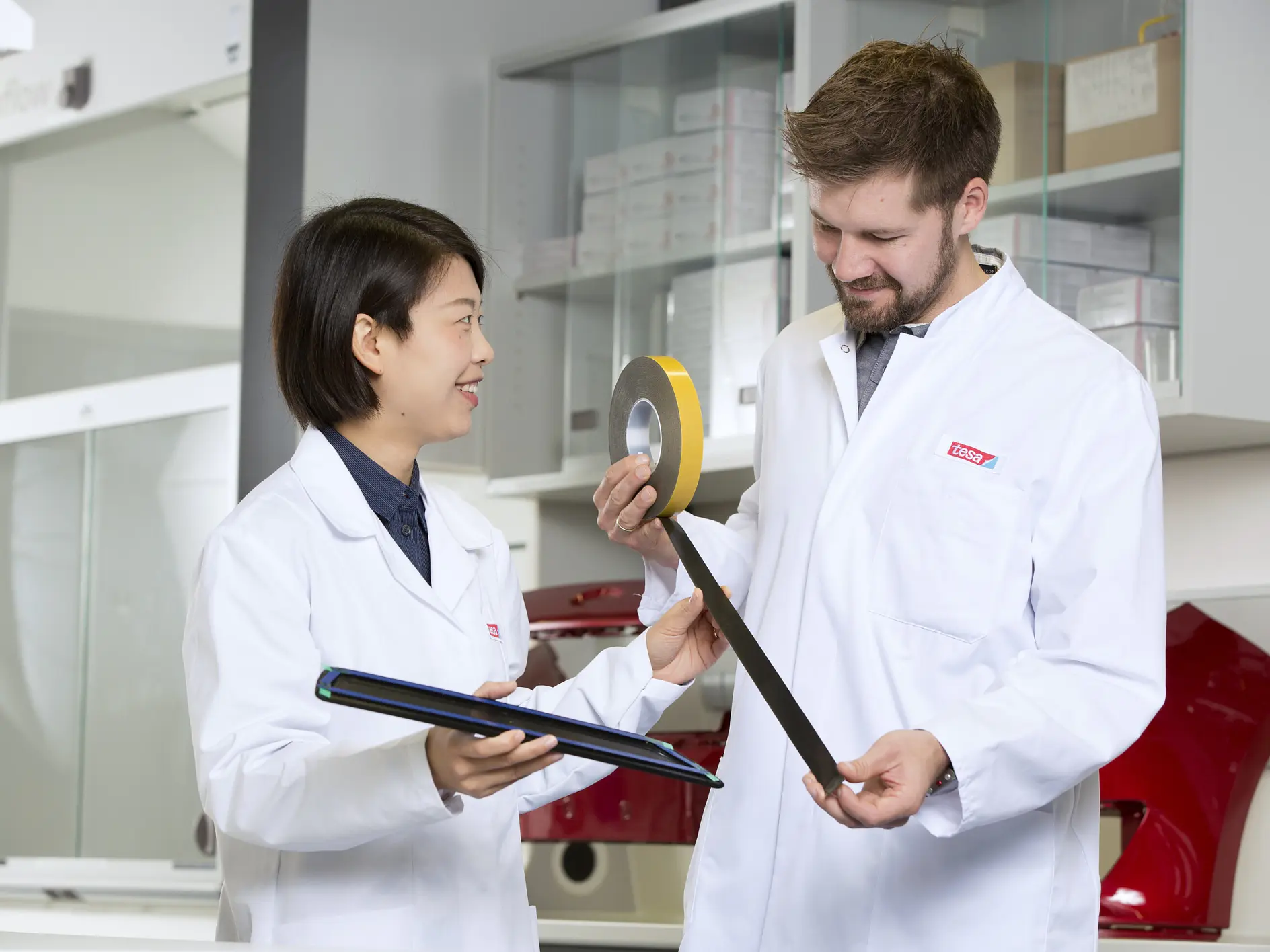 Two scientists in white lab coats discuss a black rod and yellow tape in a modern laboratory setting. (This text has been generated by AI)