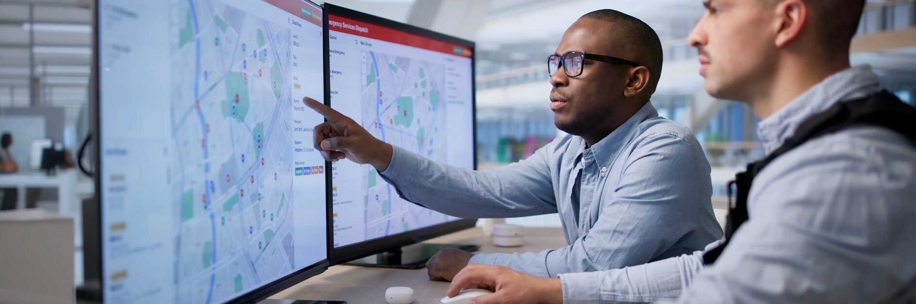 Two men sit at a desk looking at maps and data on monitors. One points at the screen. The office features tesa tape on the desk.