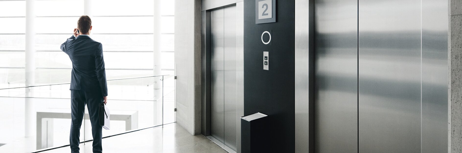 man in suit standing near two closed stainless steel elevators in bright modern office lobby with large windows tesa