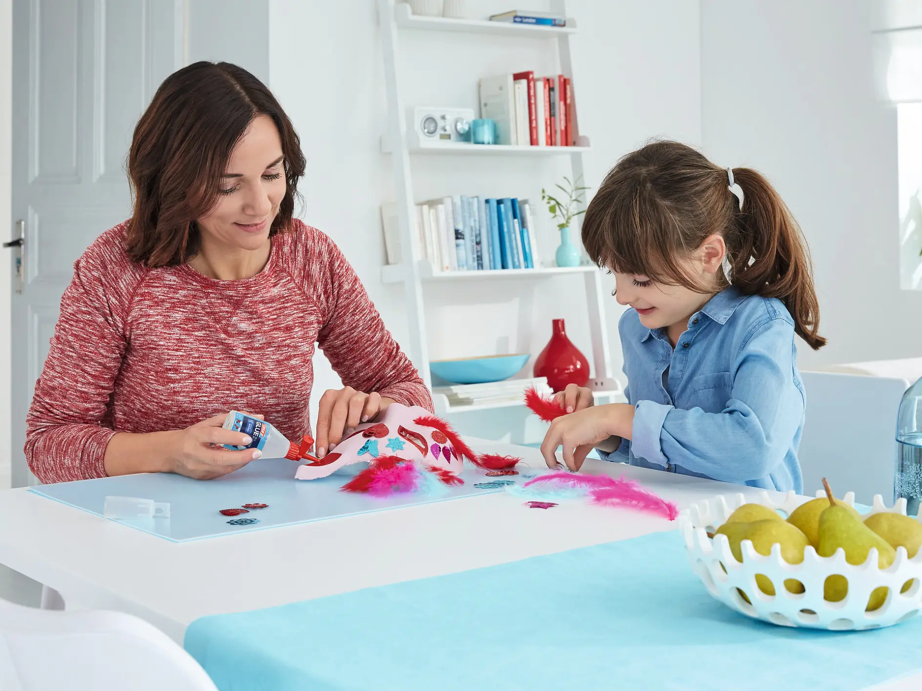 woman and young girl crafting with red paper at white table in bright room with shelves and plants (This text has been generated by AI)
