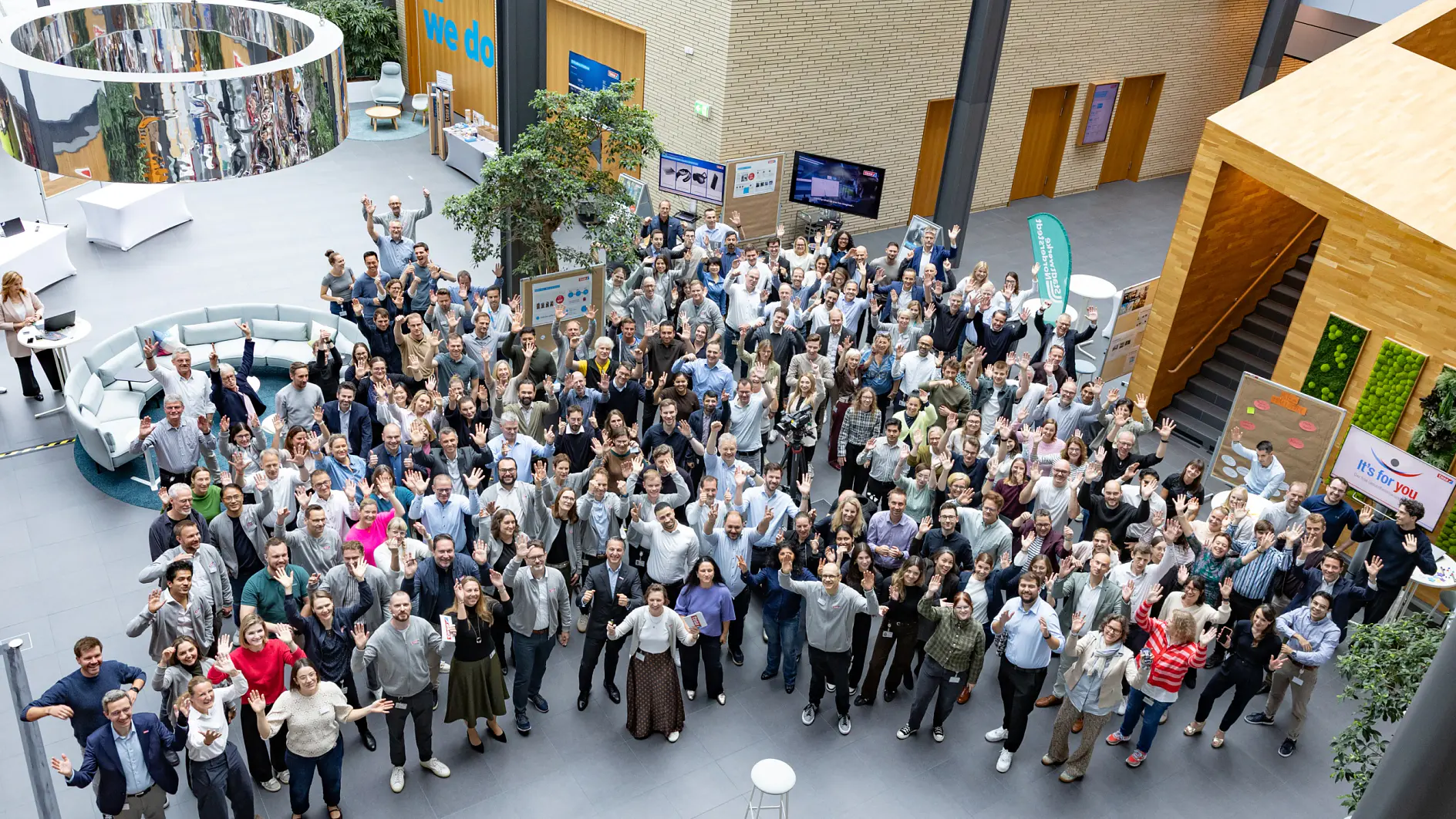 Large group of people standing and facing the camera in a modern indoor atrium, some waving and smiling. The space includes seating areas, plants, informational displays, and multiple levels with wooden and glass features. There are visible uses of tesa tape throughout the area.
