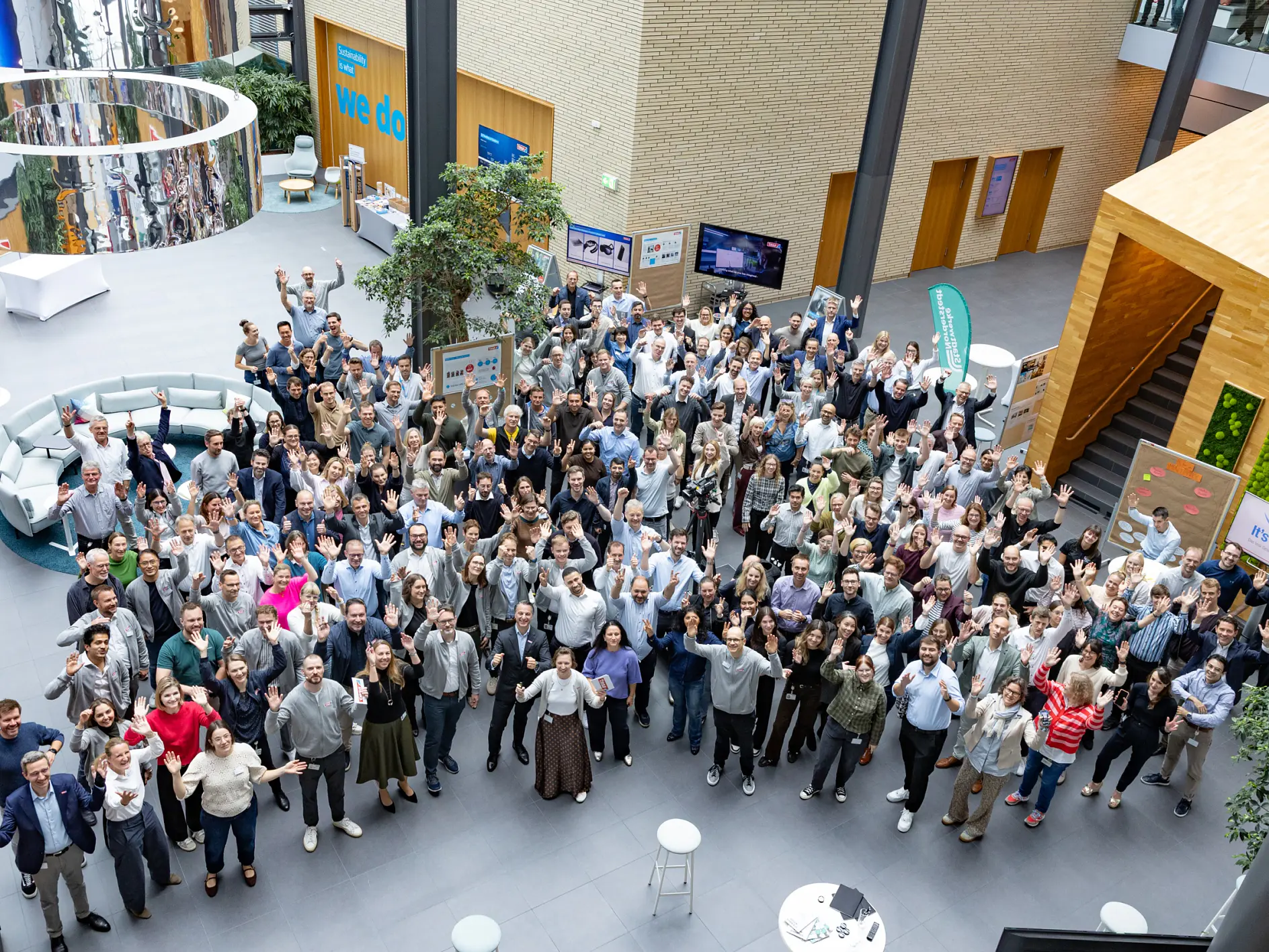 Large group of people standing and facing the camera in a modern indoor atrium, some waving and smiling. The space includes seating areas, plants, informational displays, and multiple levels with wooden and glass features. There are visible uses of tesa tape throughout the area. (This text has been generated by AI)