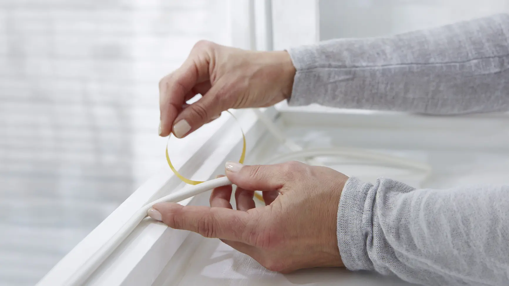 Close-up of hands applying adhesive weatherstripping tesa tape to the edge of a white window frame, likely for insulation purposes. The person is wearing a light gray long-sleeve top. Natural light comes through the window. (This text has been generated by AI)