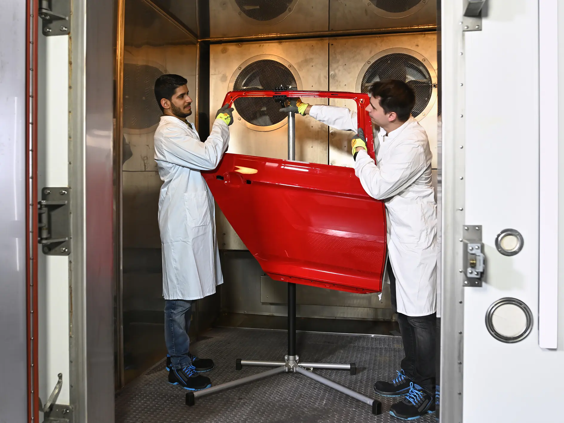 two technicians in white coats measuring a red car door inside a workshop with tools and equipment around