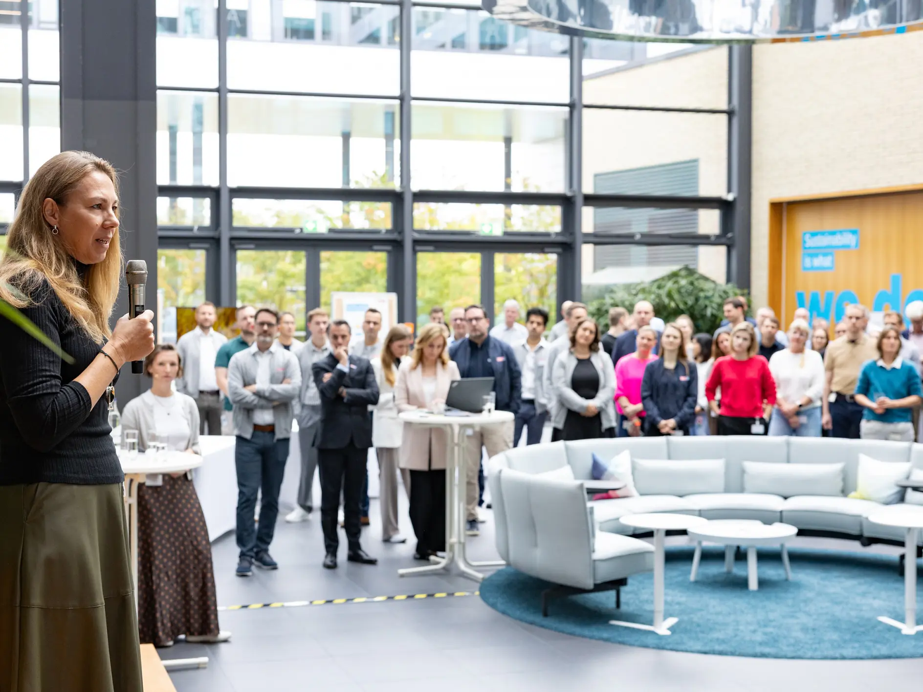 A woman stands at a podium speaking to a group of colleagues in a modern office atrium, with tesa tape visible. (This text has been generated by AI)