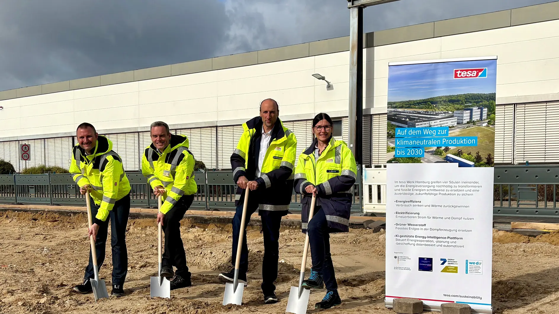 Four people in high-visibility jackets hold shovels during a ceremonial groundbreaking in front of a construction site and a tesa tape sign.