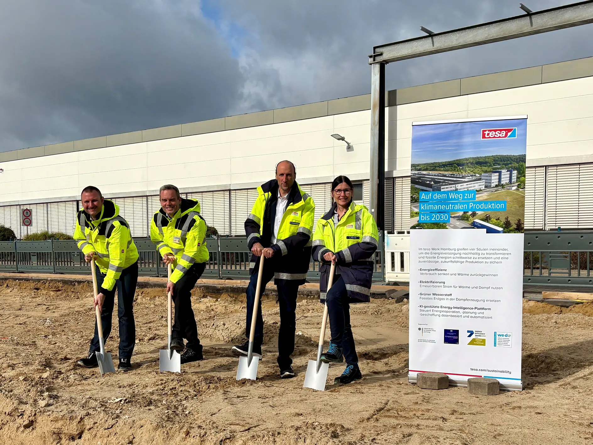 Four people in high-visibility jackets hold shovels during a ceremonial groundbreaking in front of a construction site and a tesa tape sign. (This text has been generated by AI)