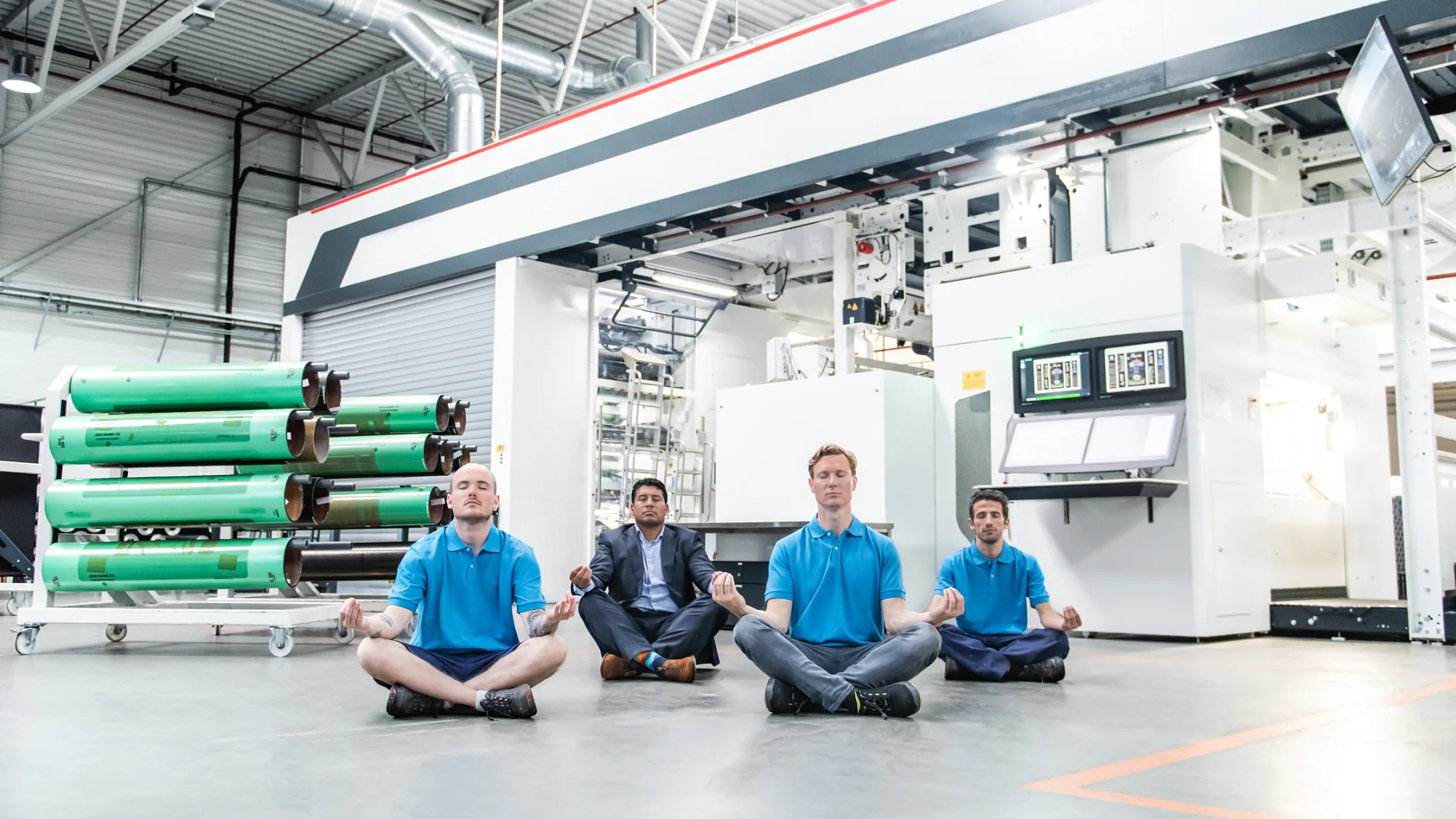 Four people sit cross-legged and meditate on the floor of an industrial workshop. They are wearing blue shirts and are positioned in front of large machinery and rolls of tesa tape material. The setting is well-lit with industrial equipment visible. (This text has been generated by AI)