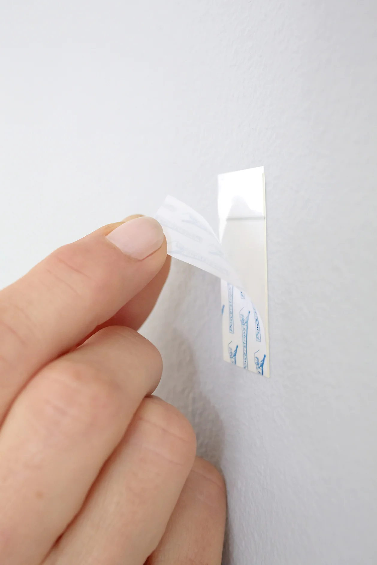 Primer plano de una mano que retira el papel protector de una tira adhesiva de pared sobre una pared de color gris claro. La tira tiene una superficie blanca con texto azul y está parcialmente adherida a la pared con cinta adhesiva tesa. Se ven las uñas de la persona. (This text has been generated by AI)