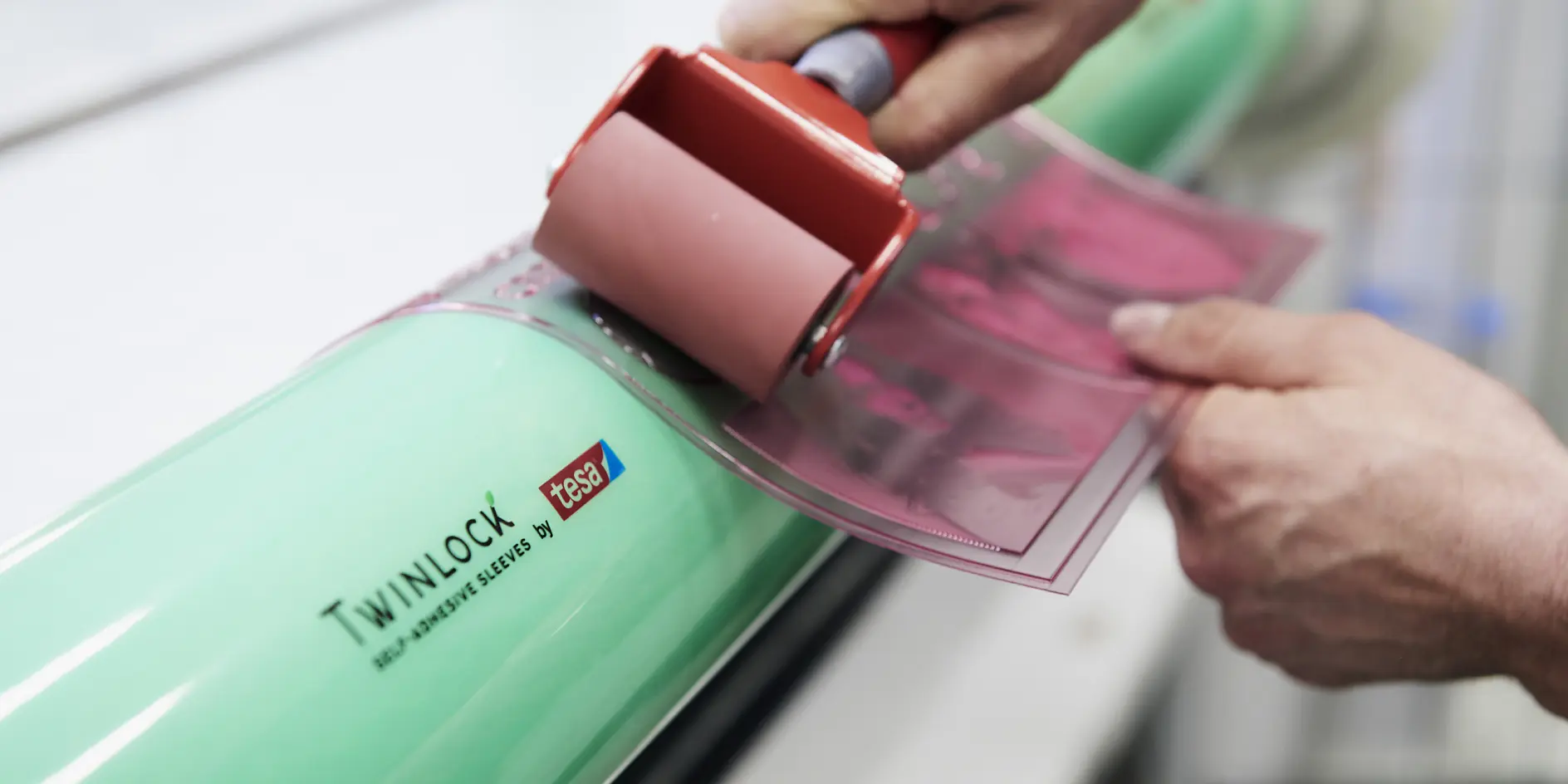 A person uses a red roller to apply a pink film onto a cylindrical green object labeled Twinlock tesa tape. The background is blurred, focusing on the hands and the application process.