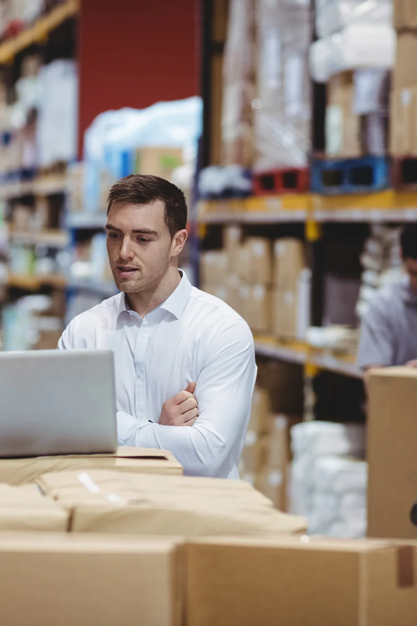 A group of warehouse workers stand by a table filled with packages. Two are engaged with a laptop, while another tallies items using a handheld device. Shelves stocked with boxes and supplies, including tesa tape, are visible in the background. (This text has been generated by AI)