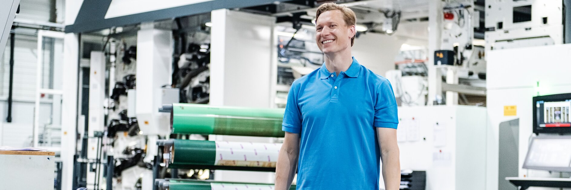 A man in a blue polo shirt and jeans is standing in a large industrial facility. Behind him, there is machinery and large rolls of tesa tape material. The setting appears to be a printing or manufacturing plant.