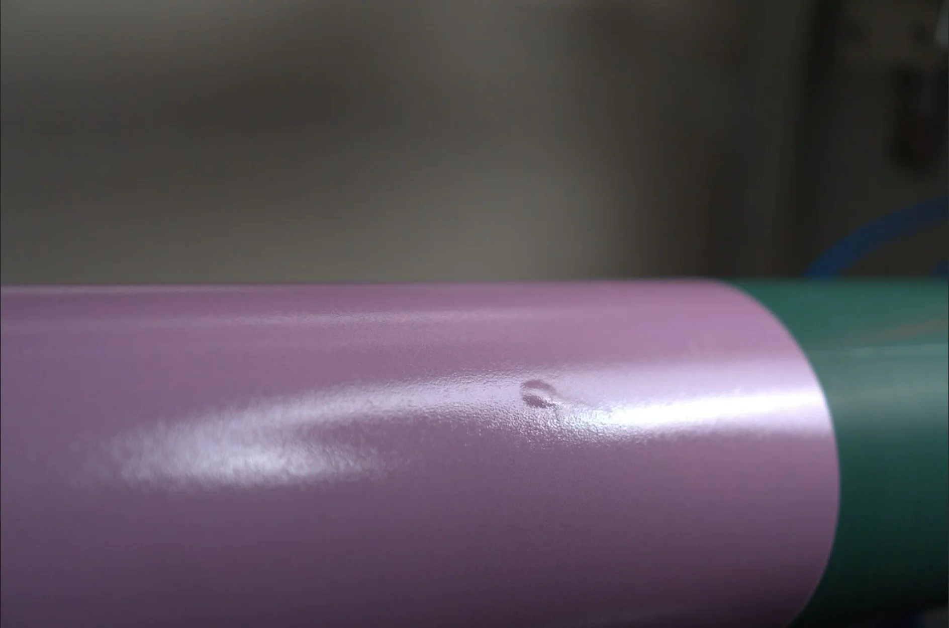 bolha-flexo Primer plano de una superficie brillante que muestra una sección violeta junto a una sección verde, con cinta tesa en uso a lo largo de la costura. El área violeta tiene una pequeña abolladura o mancha en la pintura cerca del centro de la imagen. El fondo parece desenfocado. (This text has been generated by AI)