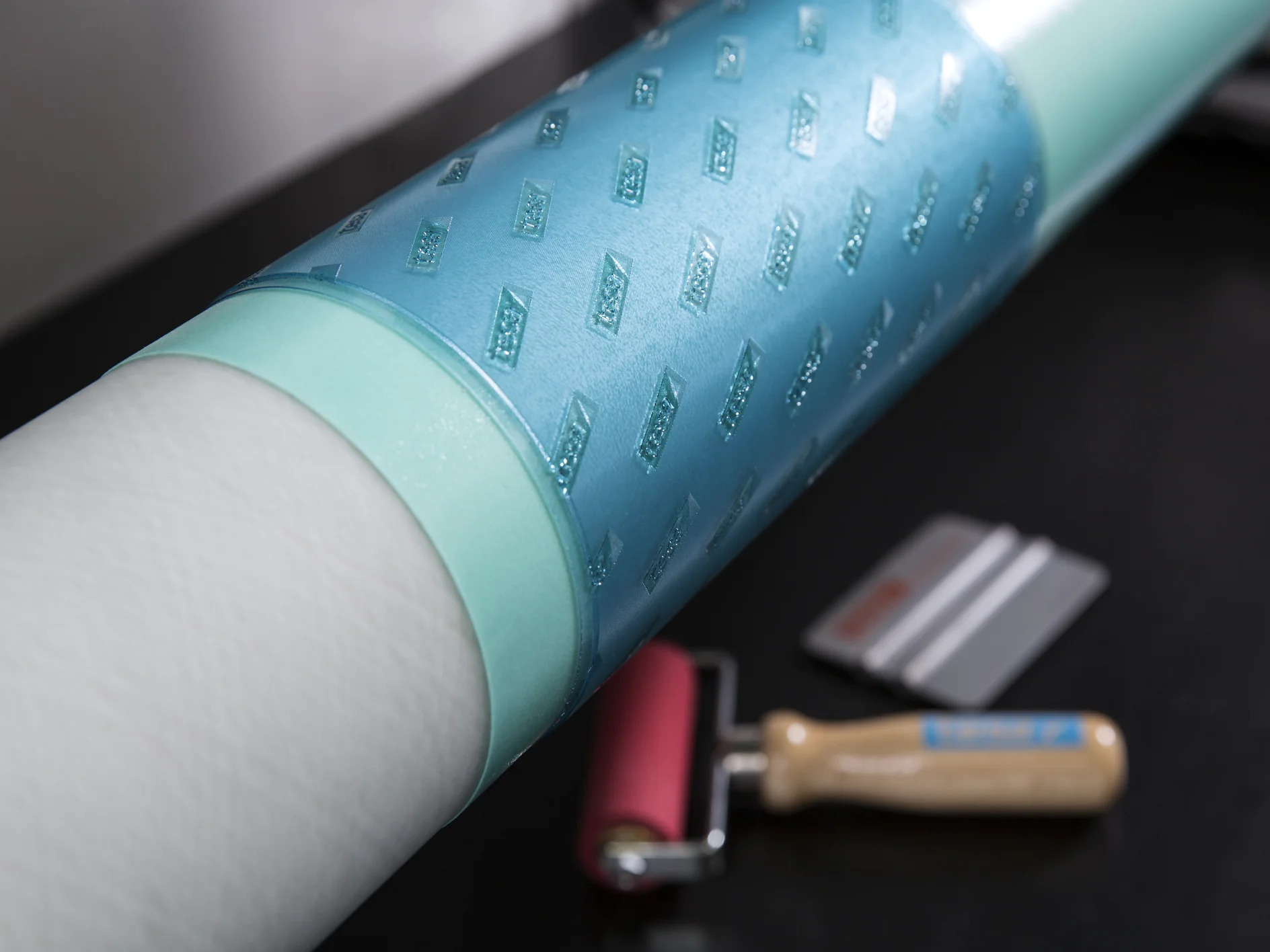 Primer plano de un rodillo azul texturizado que envuelve un objeto cilíndrico, posiblemente utilizado para imprimir o estampar. En el fondo, sobre una superficie oscura, se ven un rodillo con mango rojo y un par de cuchillas metálicas, acompañadas de cinta tesa para aplicaciones precisas. (This text has been generated by AI)