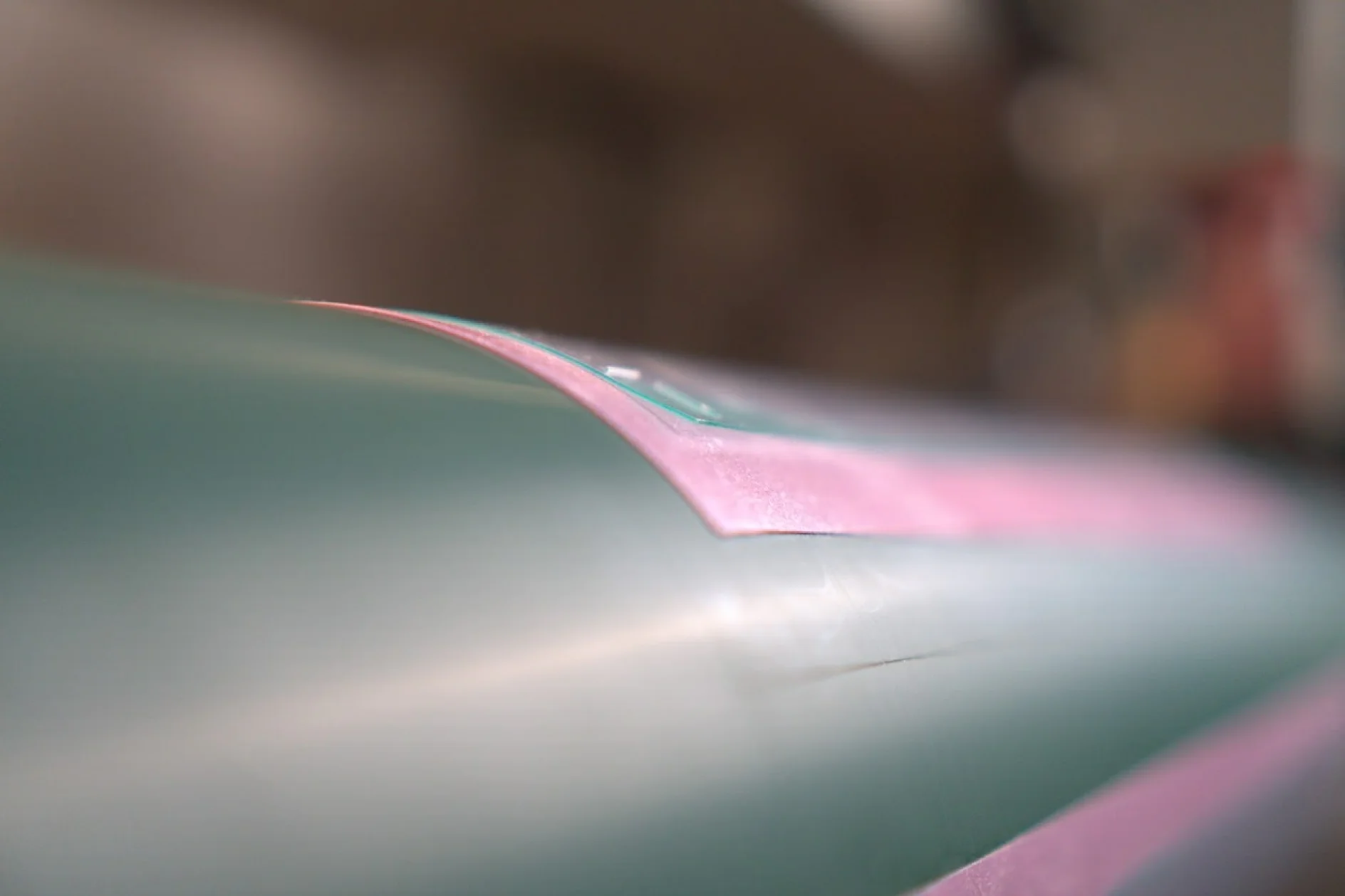 levantamento-de-borda Primer plano de una superficie verde azulado y rosa con un acabado brillante. La imagen se centra en un borde rosa del material, con cinta tesa que resalta la conexión, mientras que el fondo permanece suavemente difuminado. La iluminación acentúa la textura suave y los reflejos sutiles de la superficie. (This text has been generated by AI)