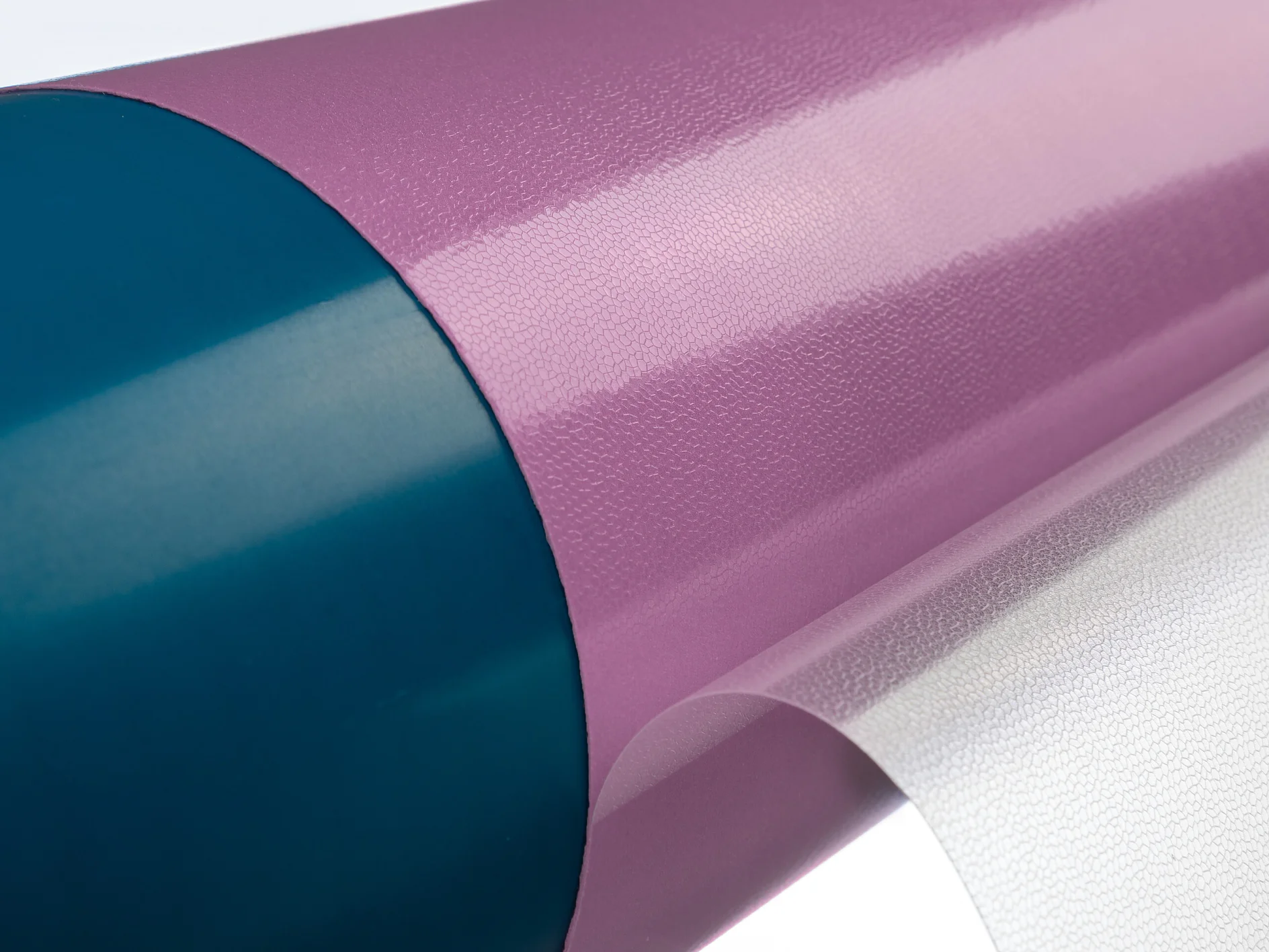 tesa-softprint-ea-flex-medium-purple-on-sleeve-with-open-liner (1) Vista en primer plano de un rollo de material de vinilo con una superficie violeta brillante asegurada con cinta tesa. El material está parcialmente desenrollado, lo que muestra su textura. El extremo del rollo es de un color verde azulado contrastante. El fondo es un degradado blanco suave. (This text has been generated by AI)
