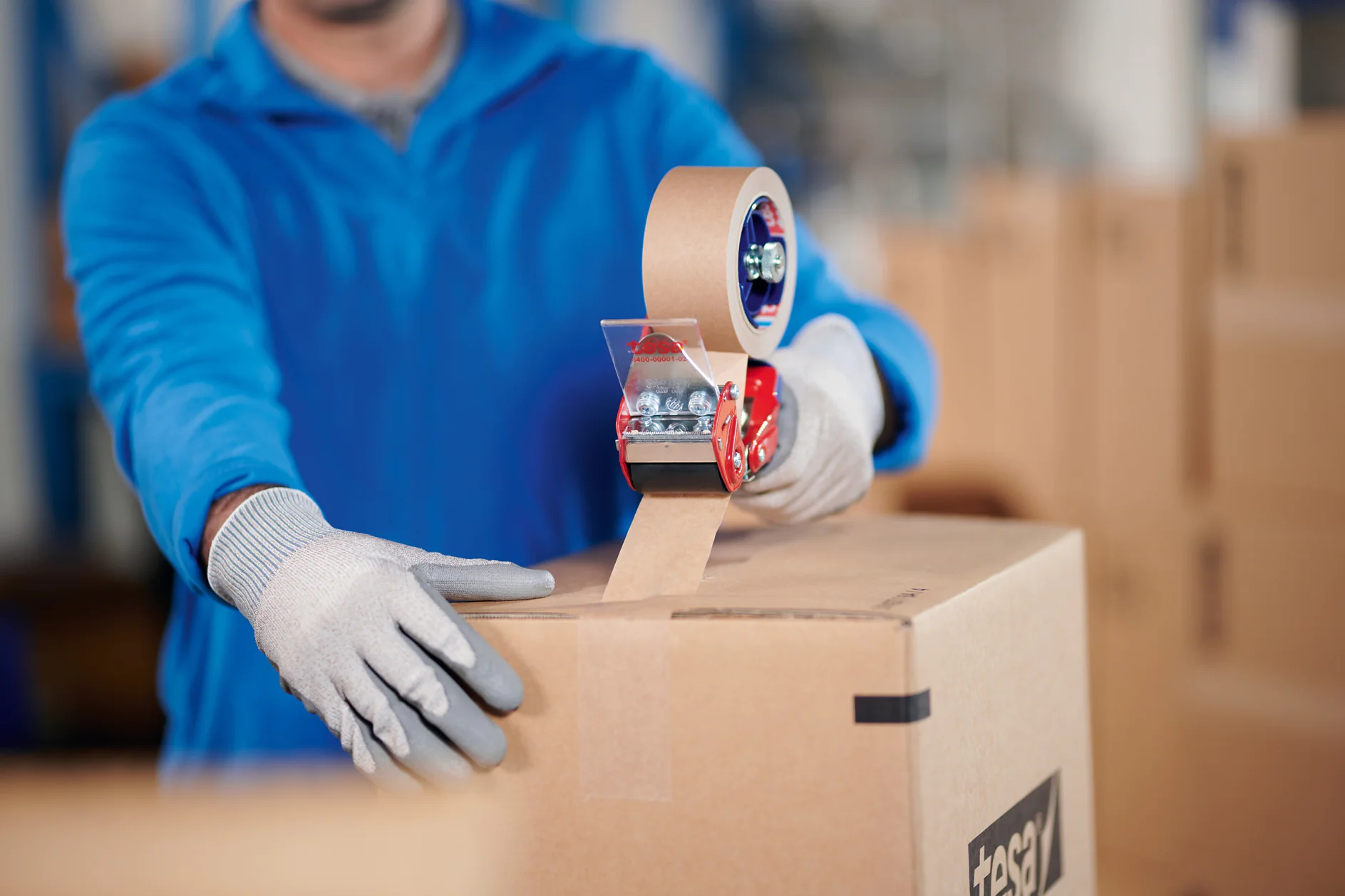 Una persona que viste una camisa azul y guantes grises está sellando una caja de cartón con cinta de embalaje Tesa de color marrón usando un dispensador manual. El fondo muestra más cajas de cartón en un entorno de almacén. (This text has been generated by AI)