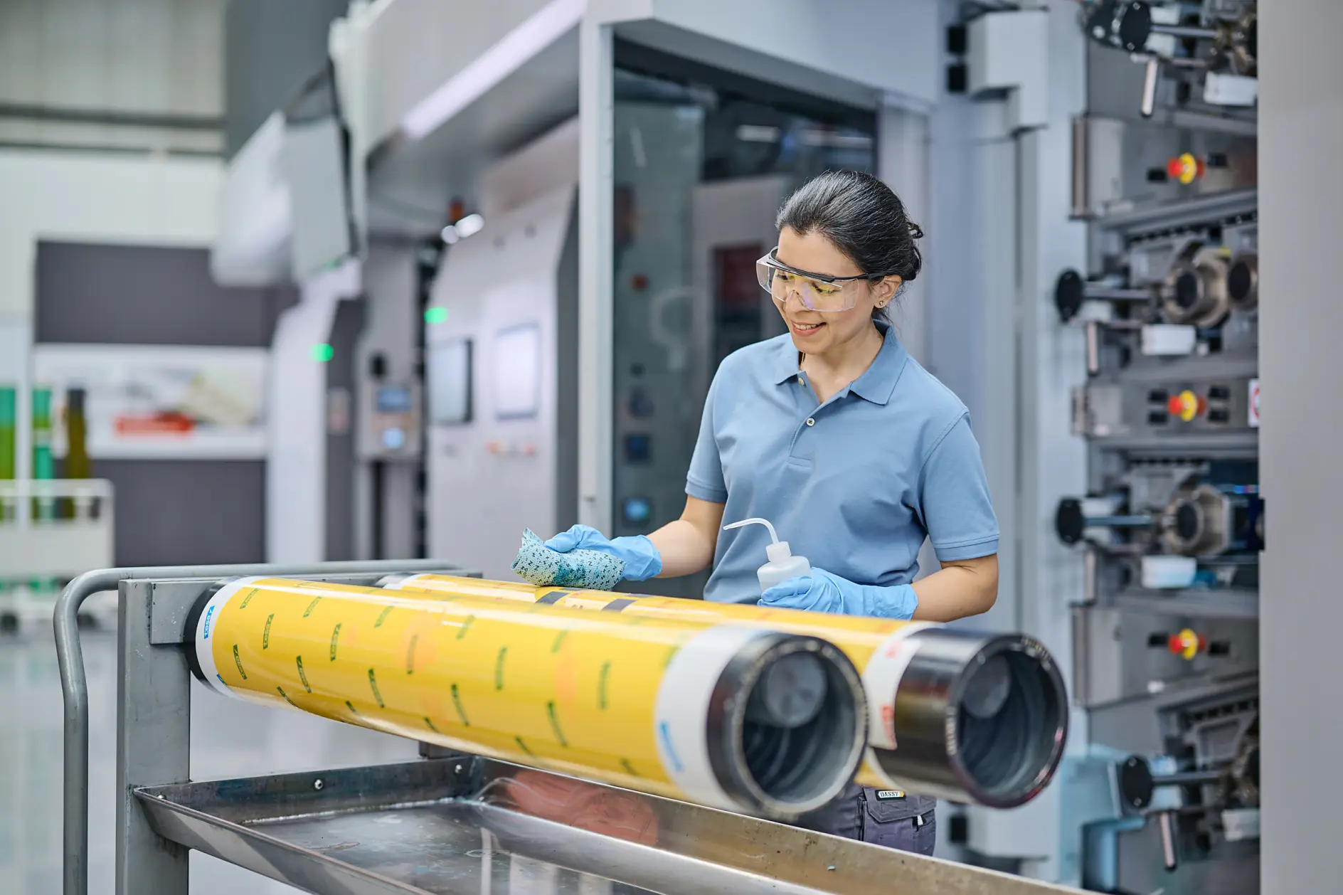 A person wearing safety glasses and gloves stands beside a printing press machine, cleaning large yellow rollers with a cloth and spray bottle. The scene appears to be in an industrial or manufacturing setting, with tesa tape nearby for additional tasks. (This text has been generated by AI)