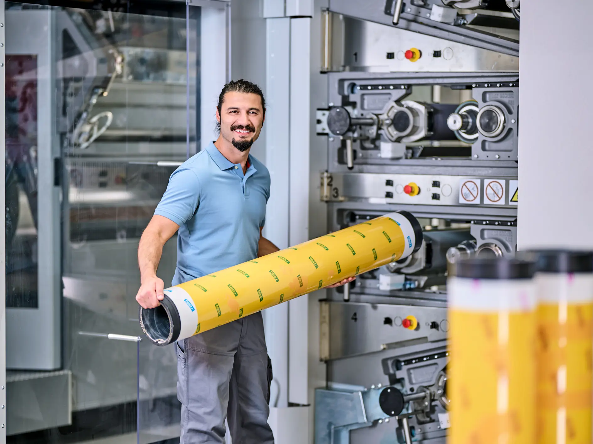 A man with a beard is holding a large cylindrical roll of tesa tape with yellow and black markings. He stands next to industrial printing machinery inside a manufacturing facility. He is wearing a light blue shirt and gray pants. (This text has been generated by AI)