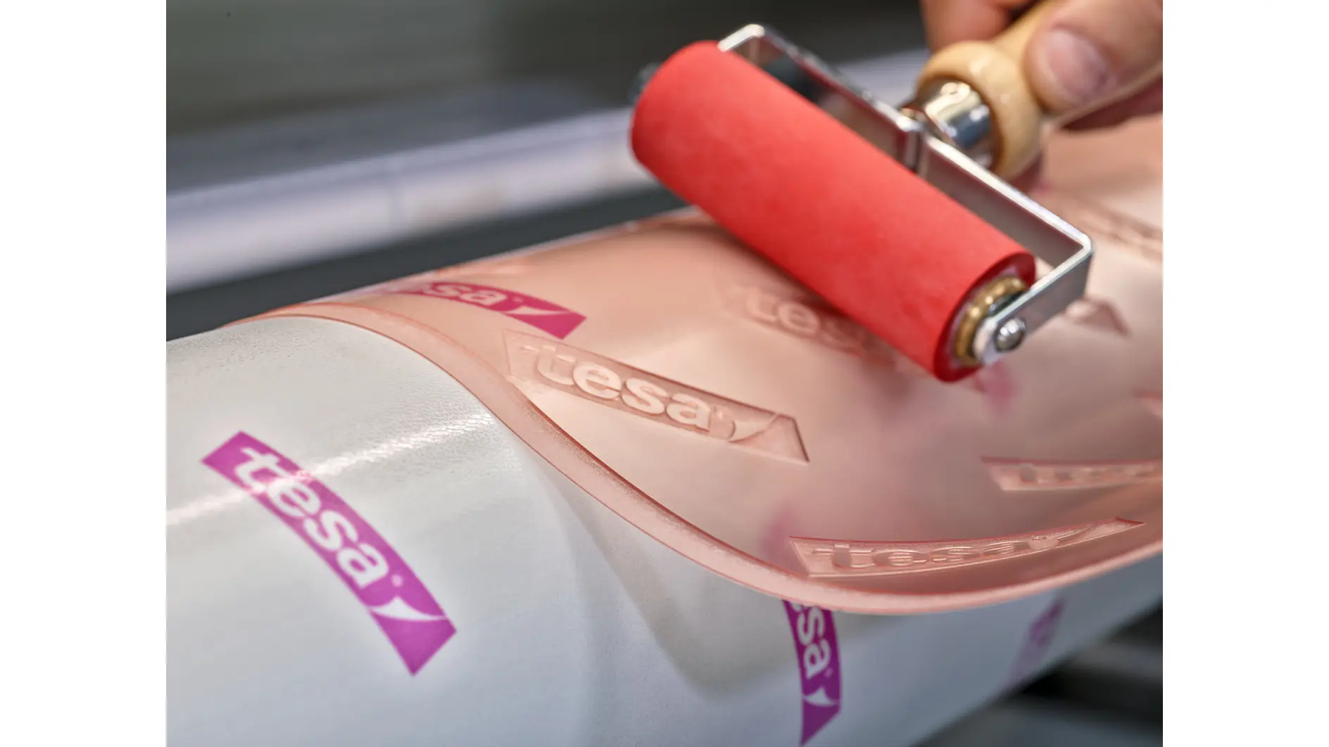 Close-up of a hand using a red roller on a clear adhesive film marked with the tesa tape logo. The film is being applied to a white cylindrical surface. (This text has been generated by AI)