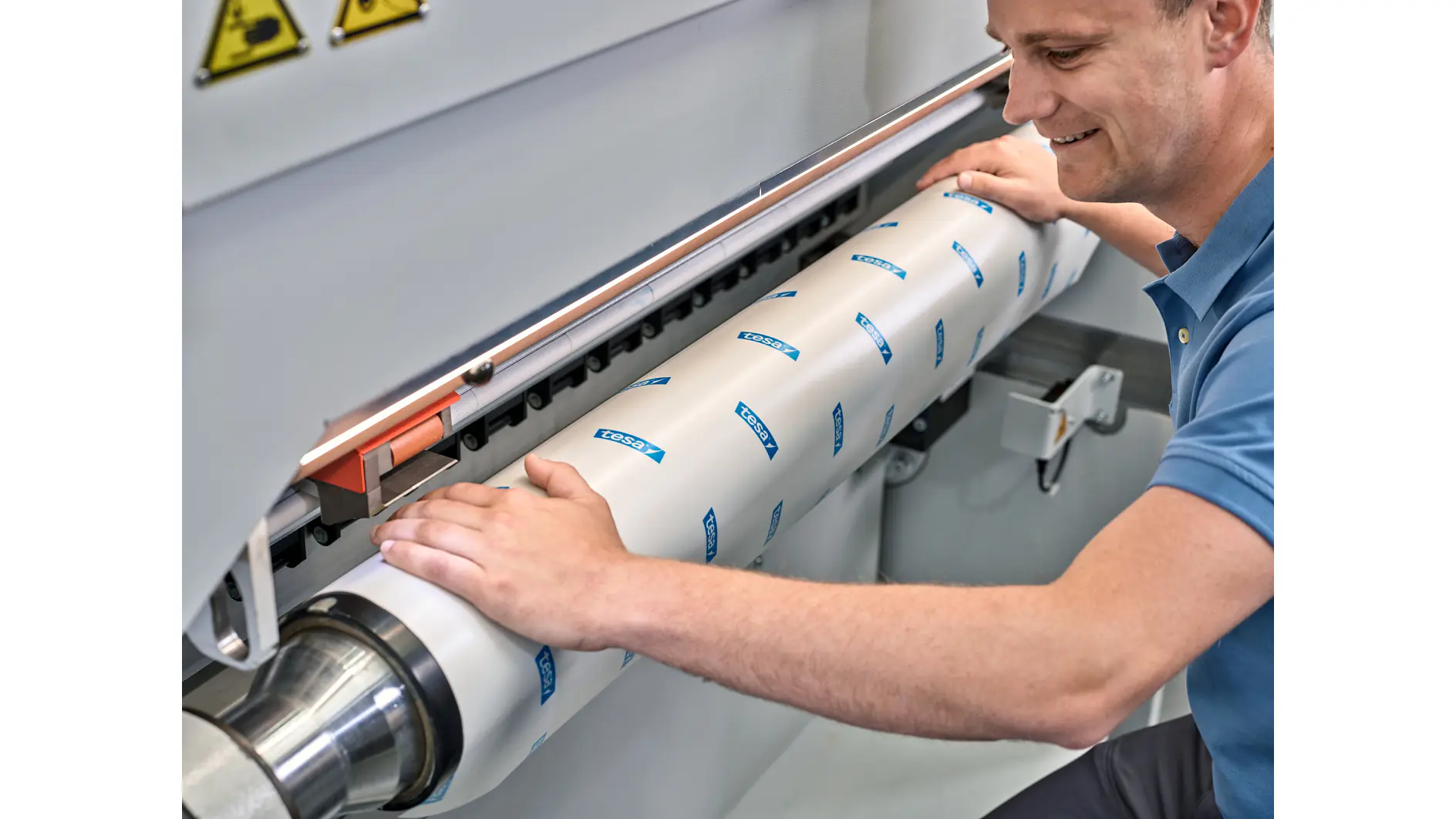 A person is operating a machine, adjusting a large cylindrical component covered with white material and blue tesa tape logos. The component is secured in place. The person is focused on the task, wearing a light blue short-sleeve shirt. (This text has been generated by AI)