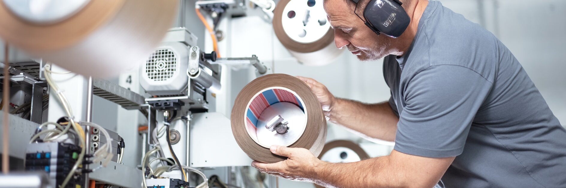 A man wearing ear protection inspects a large roll of tesa tape in an industrial setting. He is surrounded by machinery and other rolls of tesa tape, focusing intently on the one in his hands. The environment appears to be a manufacturing facility.