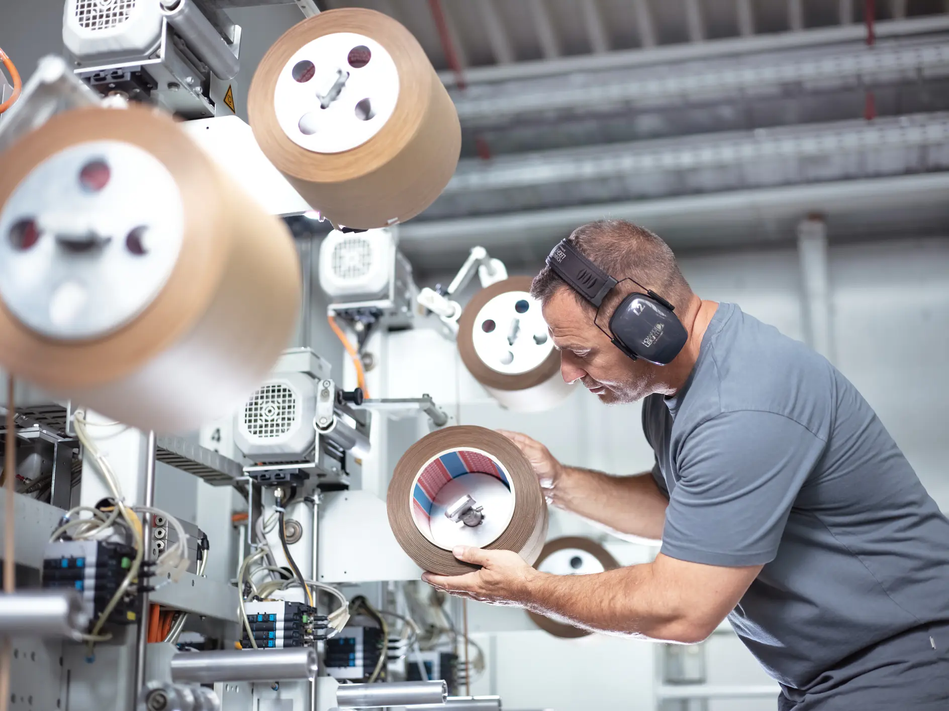 A man wearing ear protection inspects a large roll of tesa tape in an industrial setting. He is surrounded by machinery and other rolls of tesa tape, focusing intently on the one in his hands. The environment appears to be a manufacturing facility.