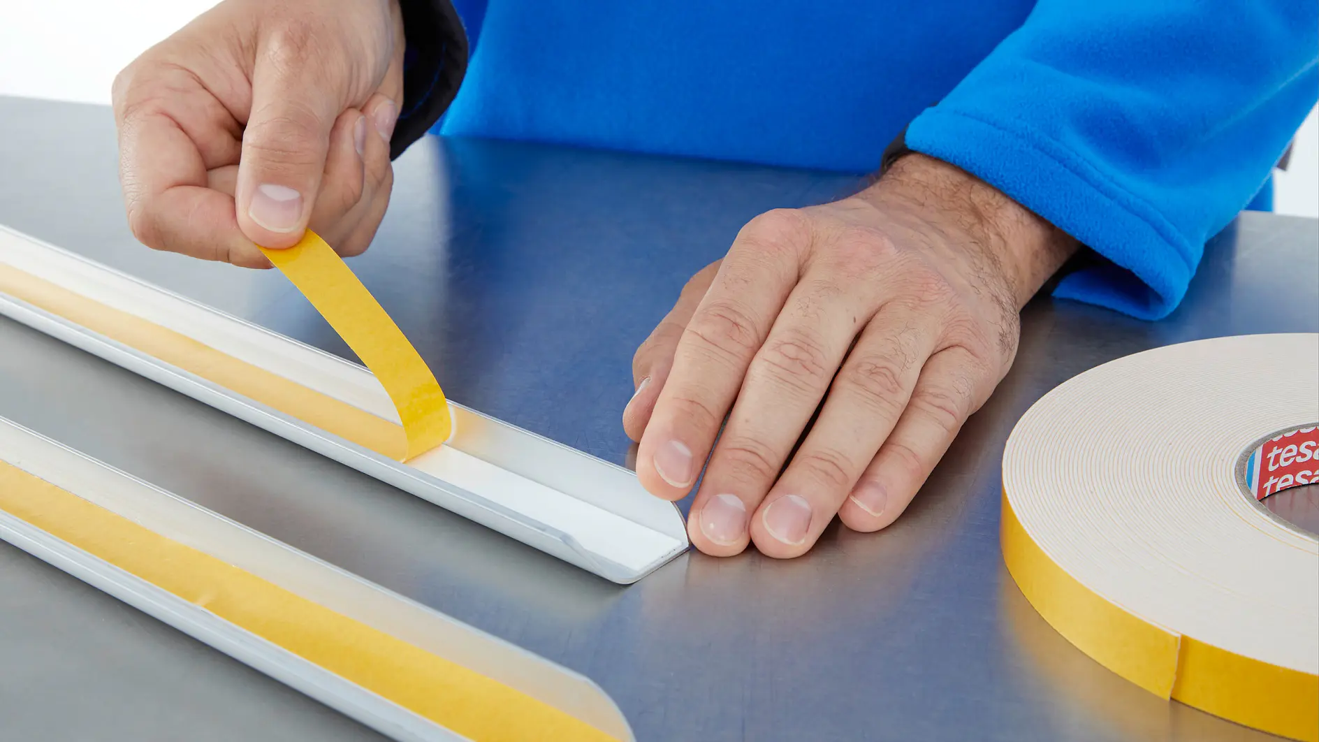 A person in a blue sleeve applies a strip of yellow double-sided tesa tape to a white rectangular object on a metallic surface. A roll of similar tesa tape is visible on the right side of the image.