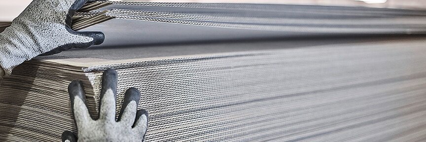 Hands wearing gloves are adjusting a large stack of corrugated cardboard sheets in a warehouse or industrial setting. The sheets are arranged in a neat pile, with strips of tesa tape securing the layers together. The background shows bright, blurred lighting. (This text has been generated by AI)