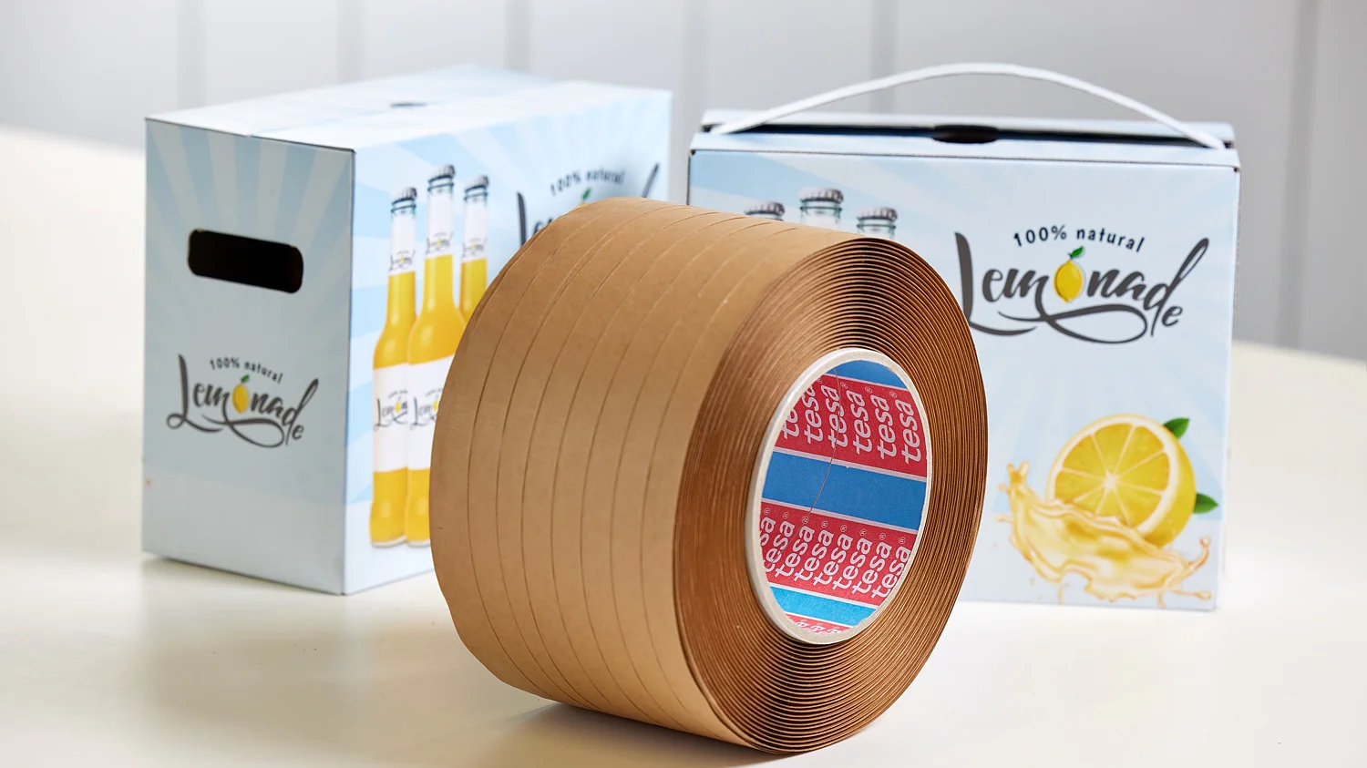 A roll of brown tesa tape is on a white surface. In the background, there are two boxes labeled 100% natural lemonade, with lemon graphics. The boxes have a handle and an illustration of lemonade bottles. (This text has been generated by AI)