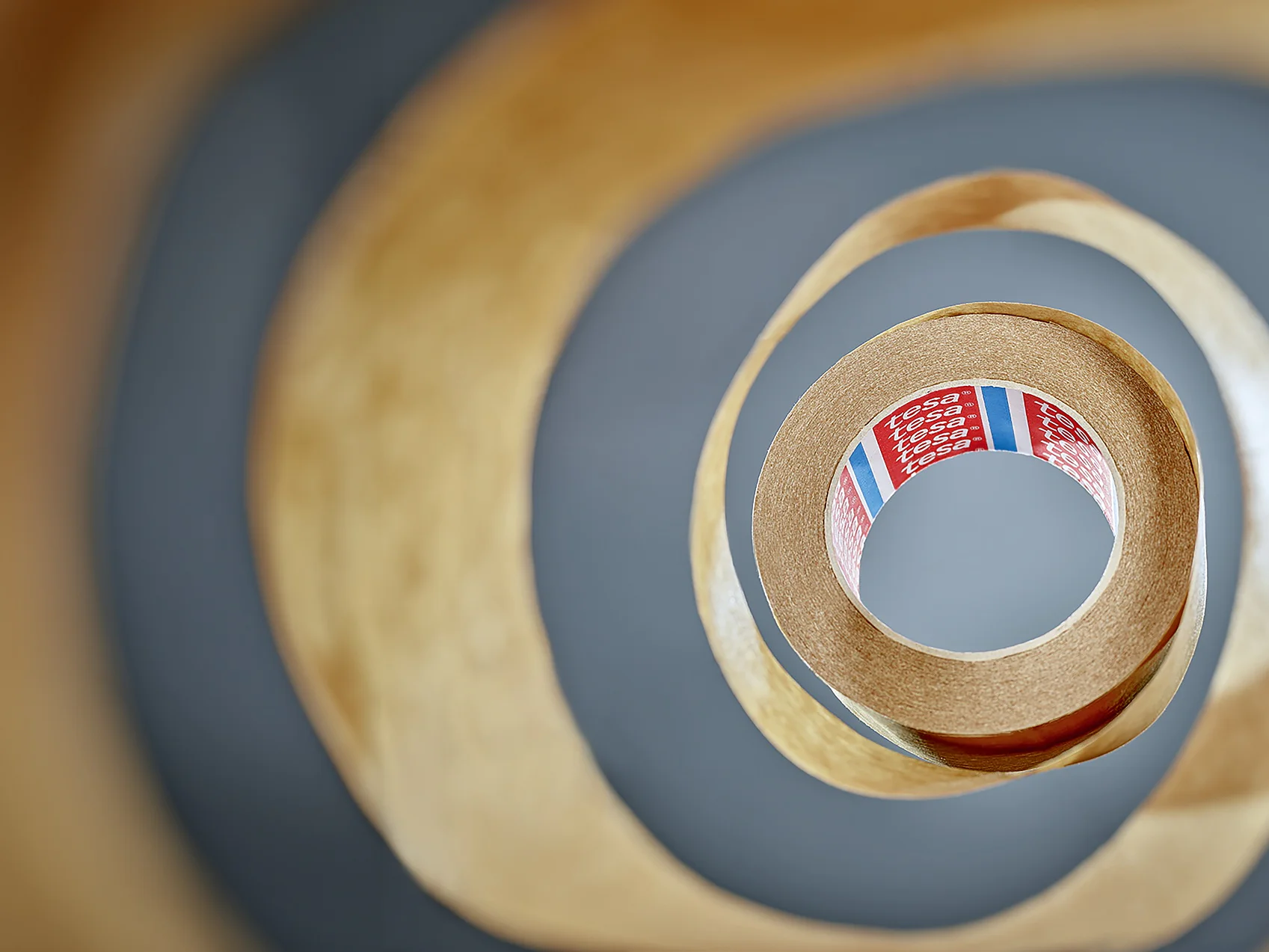 View from the center of a roll of tesa tape, creating a spiral pattern. The tesa tape is brown with a partially visible red label inside, featuring text and colored stripes. The background is blurred, emphasizing the layered effect of the tesa tape. (This text has been generated by AI)